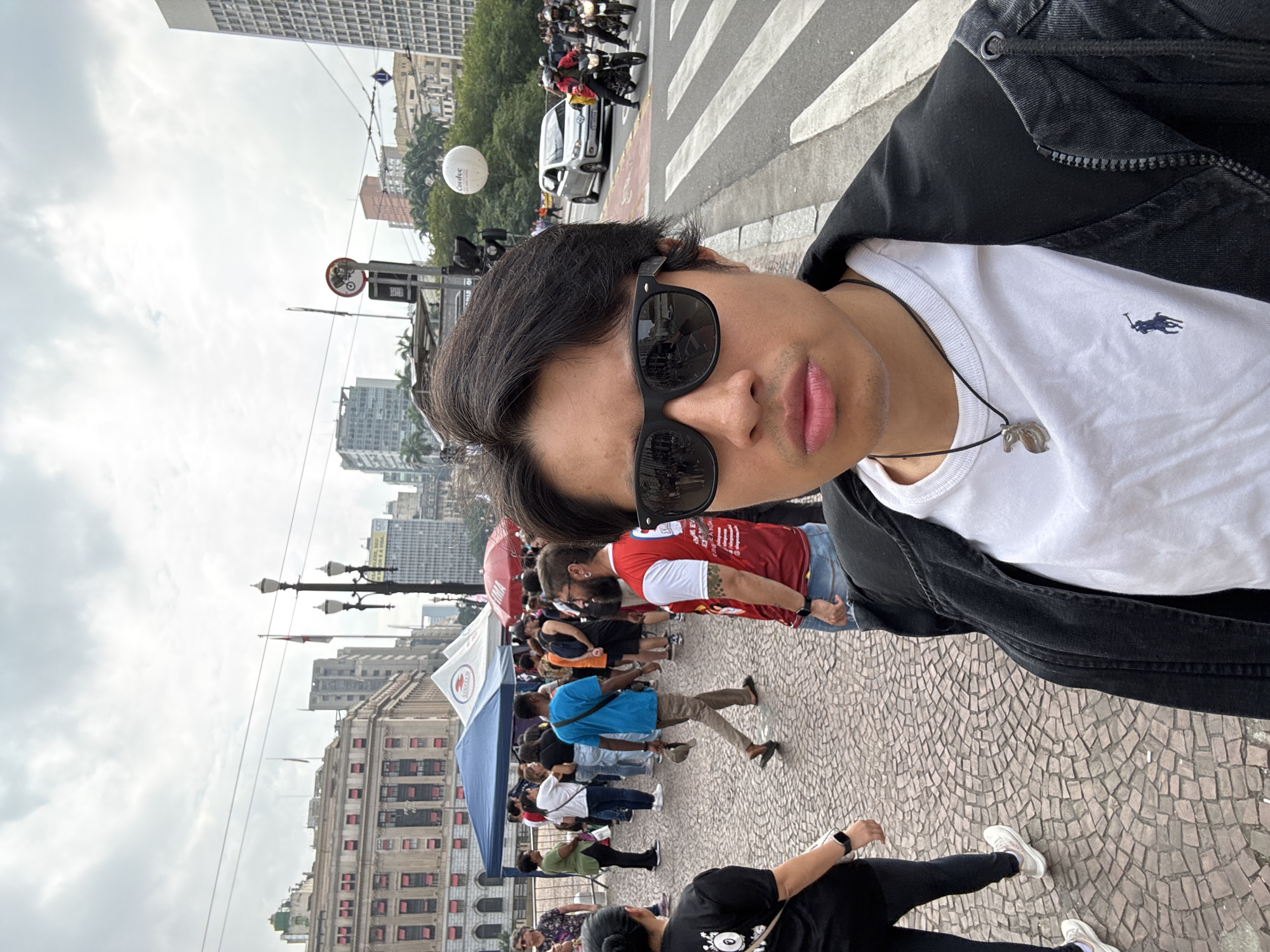 A selfie captures a person with dark hair, wearing black sunglasses, a white t-shirt featuring a blue Polo Ralph Lauren logo, and a black jacket. A necklace with a silver pendant hangs around their neck. The person has a neutral expression and slightly pink lips, and is standing on a patterned cobblestone pavement.
The scene depicts a bustling outdoor urban environment during the daytime under an overcast sky. In the midground, a street with a crosswalk is visible, where a white car and several motorcycles are present. Above the street, a white balloon floats, bearing the partially visible text "Carefree".
Further in the background, numerous tall buildings define the city skyline. On a separate cobblestone area to the right, a gathering of people is taking place around several tents, including a blue one and a red-and-white tent displaying a red cross symbol, likely indicating the Red Cross (Cruz Vermelha). Individuals are seen walking, standing, and interacting, suggesting a public event or market. Ornate lampposts and various street signs are also discernible. The overall atmosphere is that of a lively public space in a major city, consistent with São Paulo.