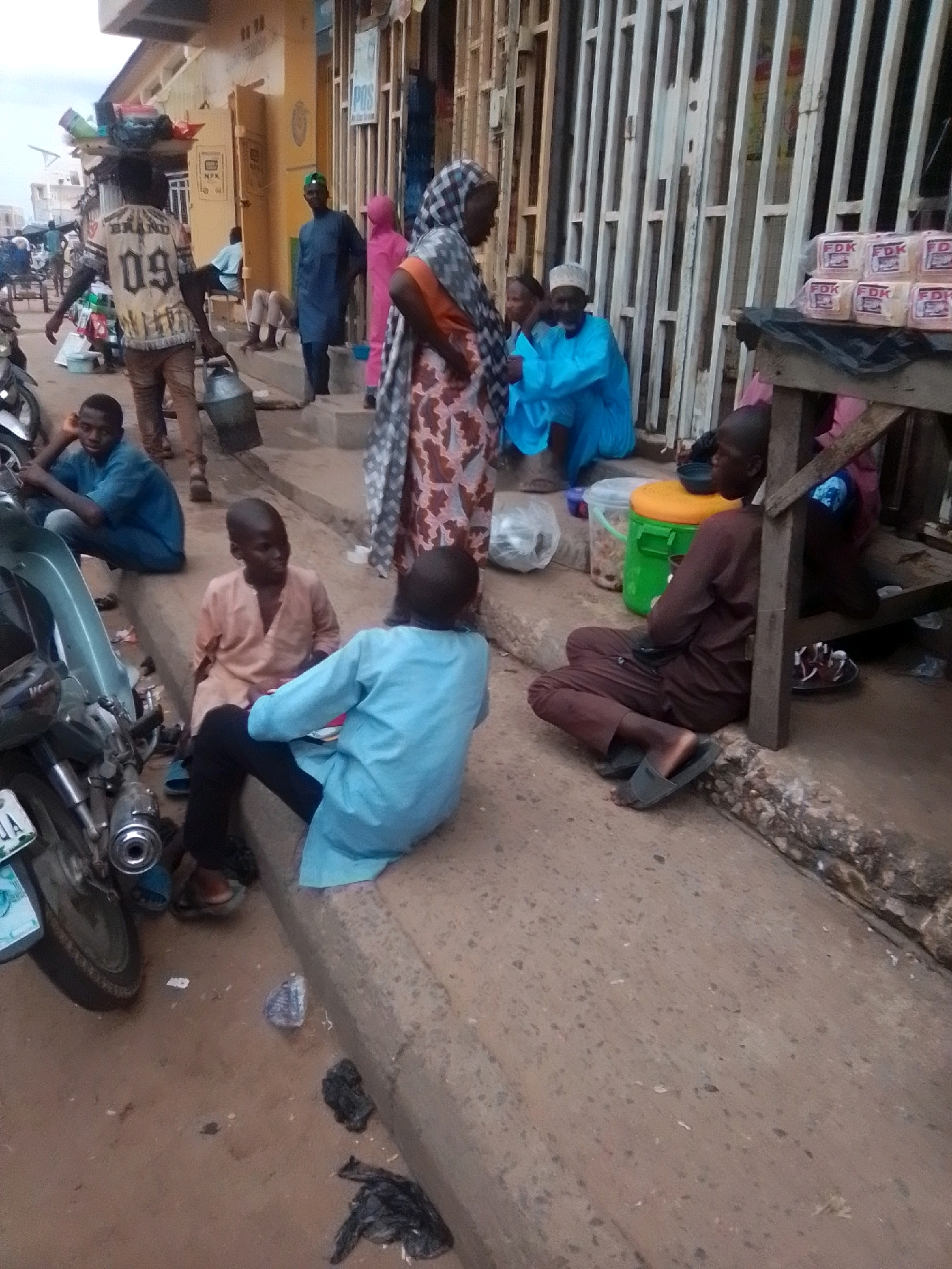 This image captures a street scene in Arawa, Nigeria, with a focus on the interactions and environment. In the foreground, a young boy in a pale peach-colored garment is seated on a curb, looking to his right. To his right, another young person in a light blue tunic and dark trousers is also seated, facing away from the camera, appearing to engage in conversation or observe something to their left. A motorcycle with a visible license plate is parked to the left of the frame.
Further back, a man with a load balanced on his head, wearing a white shirt with "BRAND 09" emblazoned on it and brown trousers, is walking, carrying a large, light-colored container. Another man in a dark blue garment and a green cap stands near a doorway. A person in a pink garment is also visible. In the background, a woman wearing a patterned headscarf and a dress is partially visible, as is a man in a blue robe sitting on the curb. A stall or makeshift table is set up, displaying stacks of bread wrapped in pink packaging with the label "FDK." Various buckets and containers are scattered around the area, including a green bucket with an orange lid. The ground is paved with concrete, and debris, including a black plastic bag, is present. The overall scene suggests a bustling marketplace or commercial area.