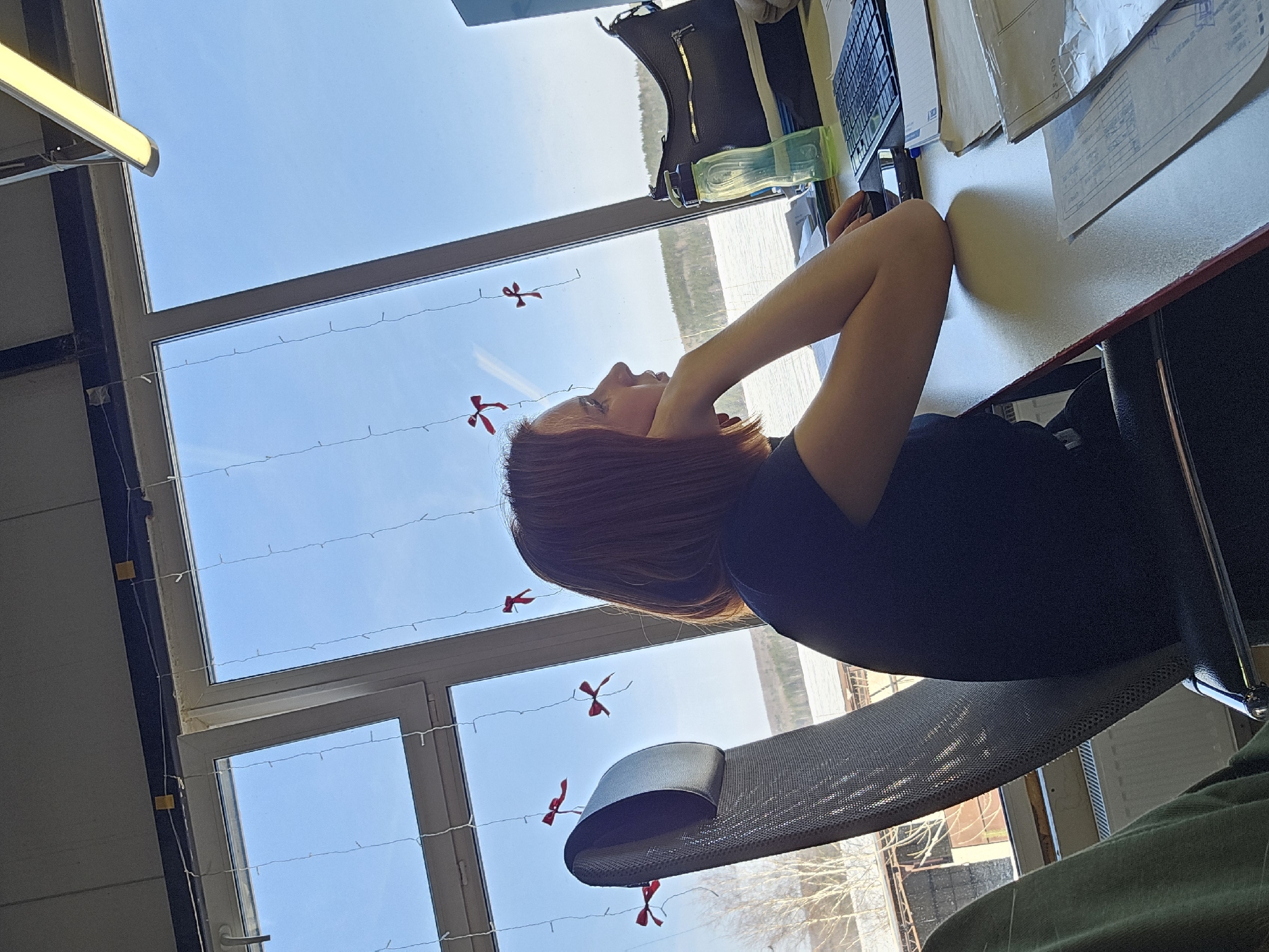 A woman with auburn hair is seated at a desk in a room with large windows in Chaikovsky, Russia. She is resting her head on her hands, looking out the window. The window has a view of a body of water and a treeline. There are decorations, possibly small red bows, strung across the window. On the desk are a keyboard, papers, a water bottle, and a handbag. The woman is sitting in a mesh office chair.