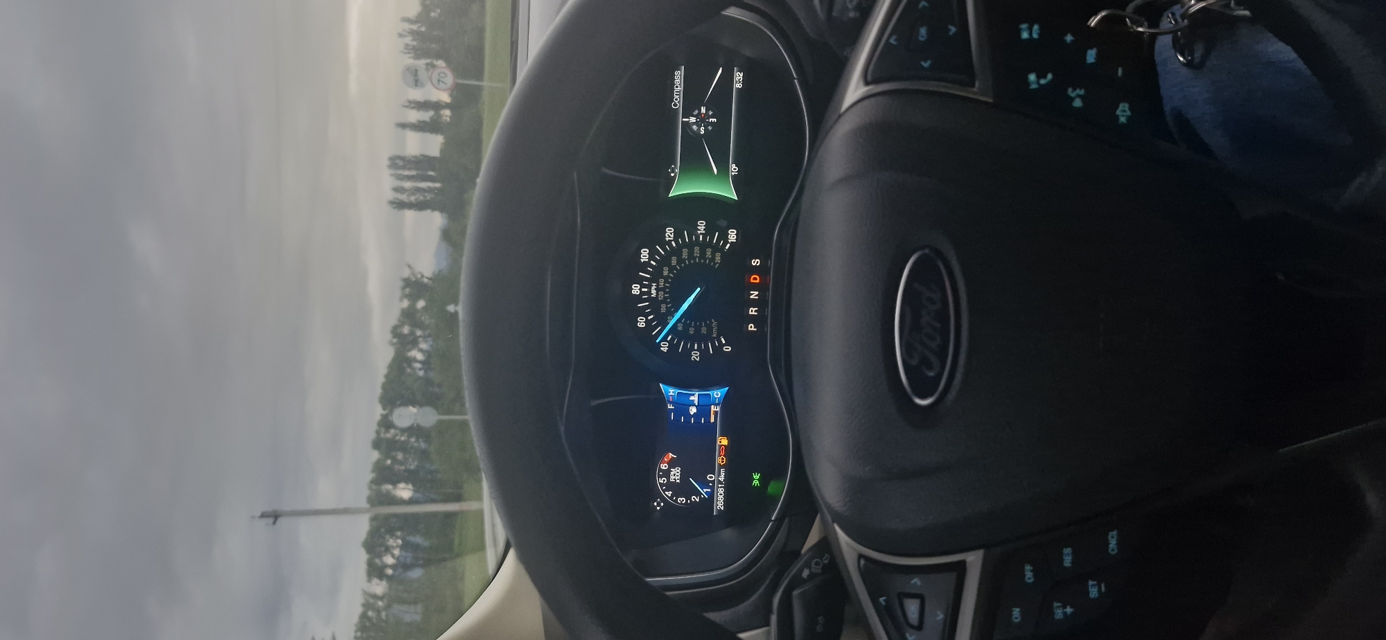The image shows the interior of a Ford vehicle, viewed from the driver's perspective. The dashboard displays the car's speed at approximately 80 mph (130 km/h), with the engine RPM at around 2000. The odometer reads 268081.4 km. The gear selector is in the 'D' position, indicating drive. A compass is visible on the right side of the dashboard, showing a temperature of 10 degrees Celsius and a time of 8:32. The exterior view shows a cloudy sky, trees lining the road, and a street light. Traffic signs are visible in the distance. Based on the presence of road signs and the overall landscape, the image is likely taken on a road in or near the city of Kutaisi, Georgia. There are no people visible in the frame. The steering wheel has controls for the cruise control system.