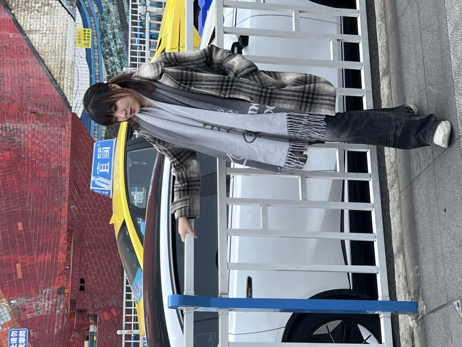 A young woman stands in front of a yellow taxi and a white car, holding onto a railing. She is wearing a plaid jacket, a gray scarf, and dark pants with white shoes. The background includes a red brick wall and a colorful mosaic pattern above the taxi. The photo is taken at an angle, with the woman positioned slightly off-center.
