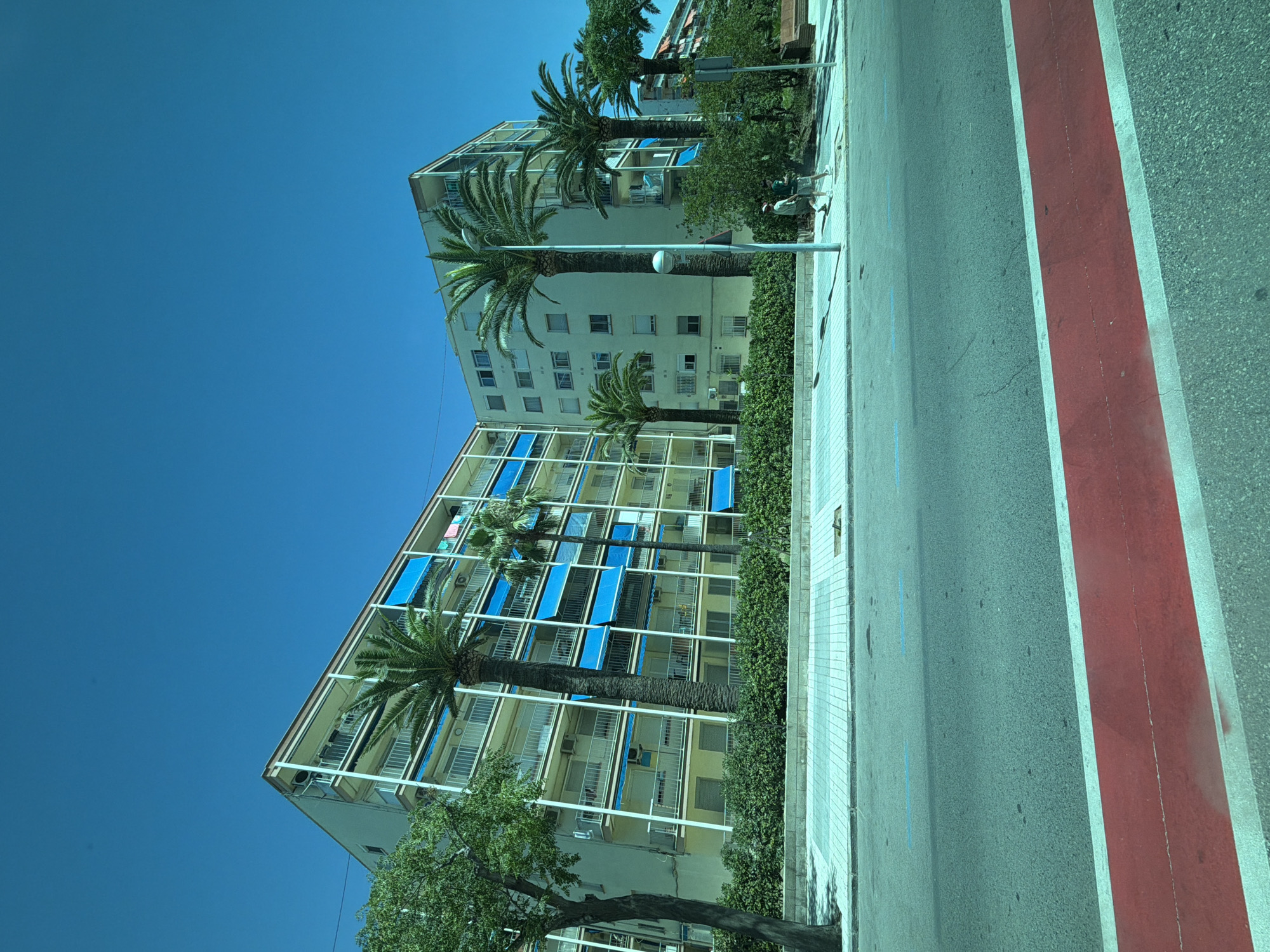 In La Pineda, Spain, a sun-drenched street scene unfolds with a clear blue sky overhead. In the mid-ground, two multi-story apartment buildings stand side-by-side. The building on the left is a light yellow, while the one on the right is a pale cream color. Both structures feature numerous balconies with white railings, many adorned with vibrant blue retractable awnings. Tall palm trees are strategically planted in front of the buildings, their fronds reaching towards the sky. A dense green hedge lines the base of the buildings.
The foreground is dominated by a street with a prominent red lane painted across it, flanked by white lines. Further back, a paved sidewalk is visible, distinguished by a light grey, geometric tile pattern. Two individuals, appearing to be young men, walk along the sidewalk away from the camera. One is wearing a light-colored t-shirt and shorts, while the other is in a darker t-shirt and shorts. A single lamppost stands on the sidewalk between the buildings and the road. The overall impression is one of a warm, sunny day in a coastal urban environment.