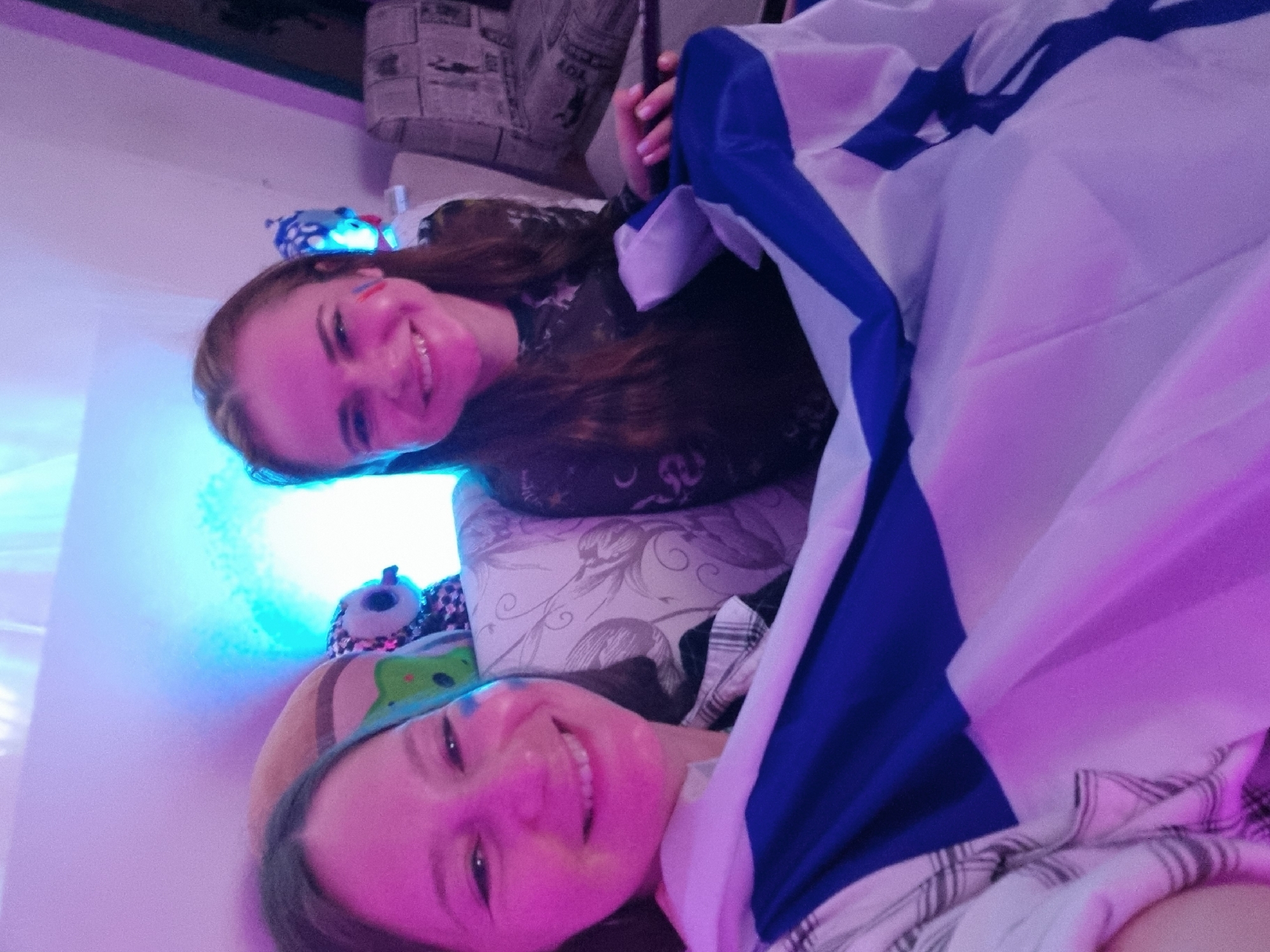 Two smiling women are lying down, covered with an Israeli flag, in an indoor setting. The setting includes a pillow and a couch with a newspaper pattern. The location is Bat Yam, Israel. One woman has blue and white paint on her cheek. The lighting in the room is a mixture of blue and purple. There are stuffed animals visible in the background.
