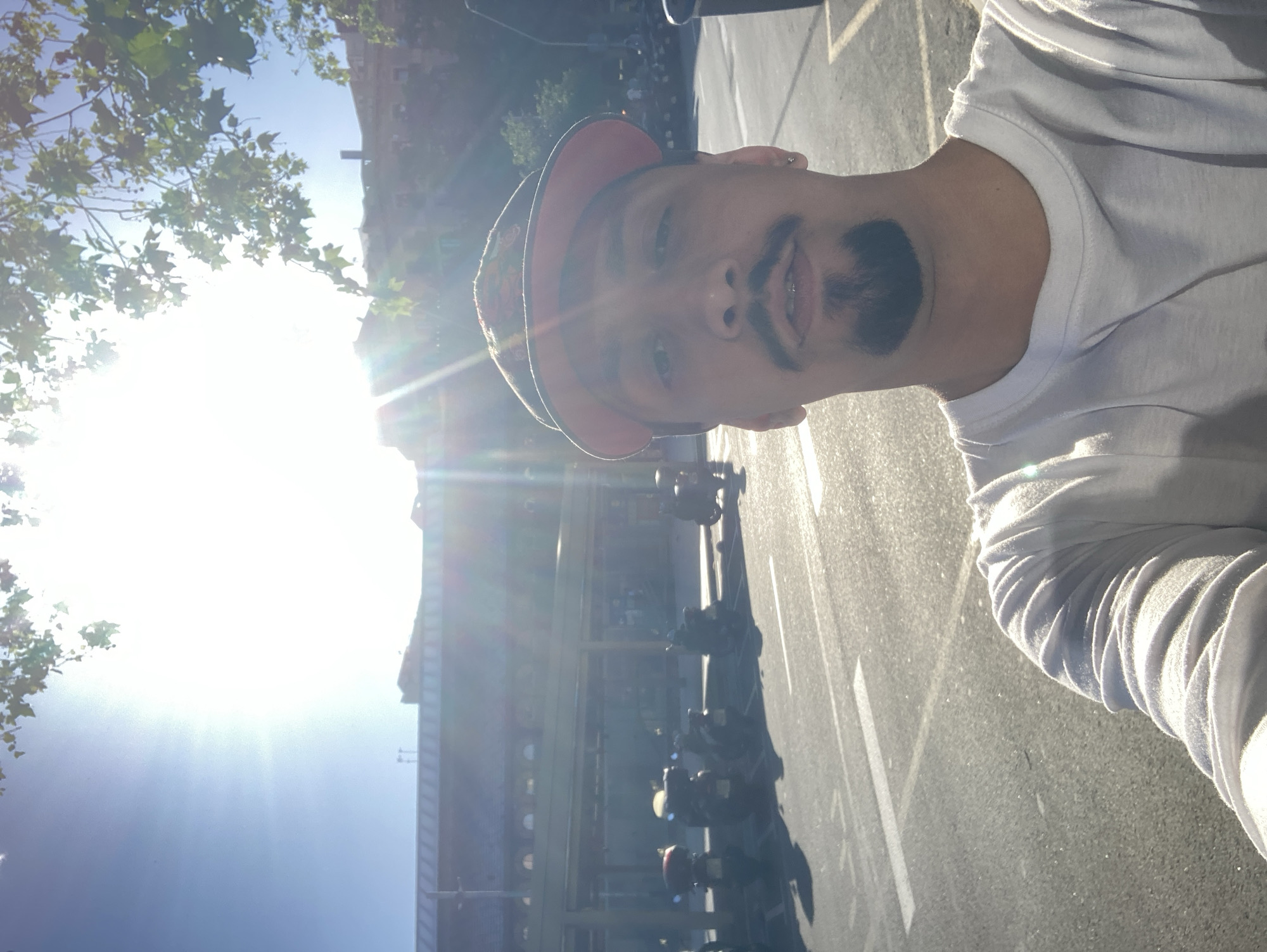 A man is standing on a city street on a sunny day. He is wearing a white t-shirt and a baseball cap. A building is in the background, and there are motorbikes parked along the street.