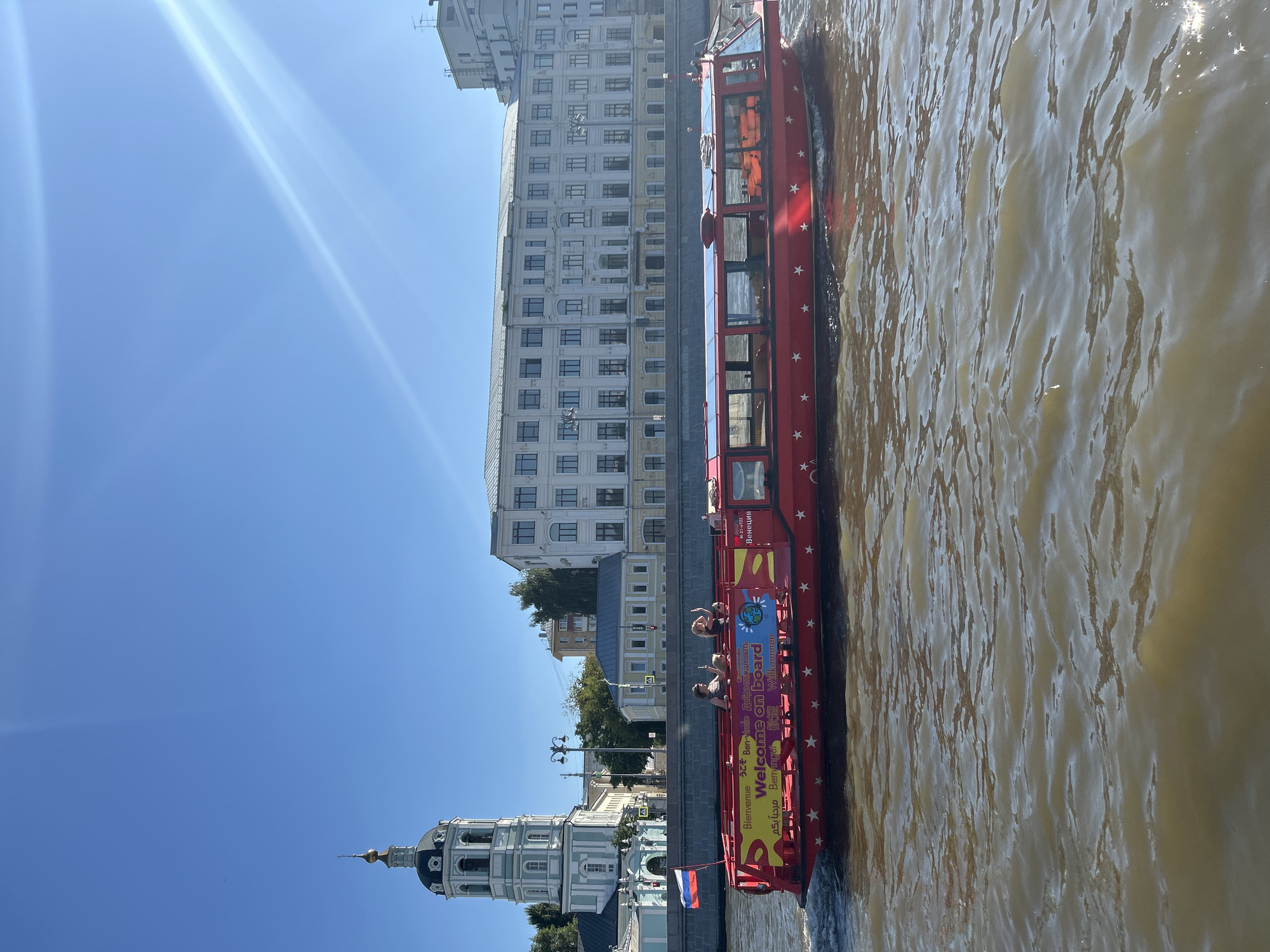 The image depicts a red tour boat cruising on a river in Moscow, Russia, under a clear blue sky with bright sun glare. The boat is decorated with white stars and features signage with multilingual greetings, including "Welcome on board." Several passengers are visible on the open deck of the boat, with two individuals appearing to wave.
In the background, a light blue church with a black dome and a golden spire stands prominently on the left. Adjacent to the church, a long, multi-story building with many windows is visible. Further in the distance, other buildings and trees line the riverbank. A Russian flag is attached to a flagpole on the embankment. The river water is a muddy brown color, with ripples and reflections of the sunlight. The overall scene captures a sunny day in Moscow, likely during a tourist season, with a river cruise in progress.