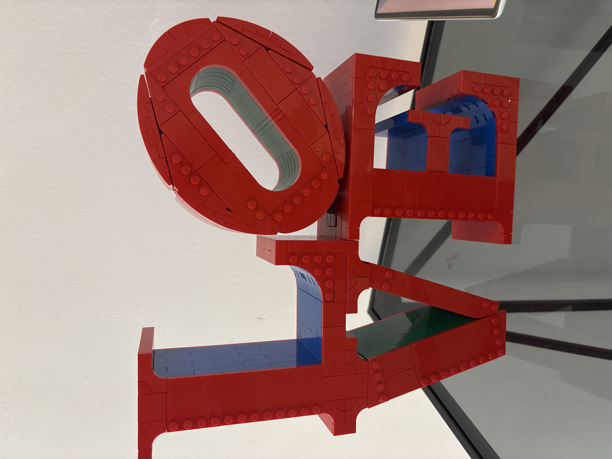 The image shows a LEGO sculpture of the word "LOVE" on a glass-top table. The sculpture is red with blue and green accents visible inside the letters. The background is a plain white wall. There are no people or animals in the image, nor any specific geographical features that would indicate its location in Dubai, United Arab Emirates. The object is a decorative art piece.