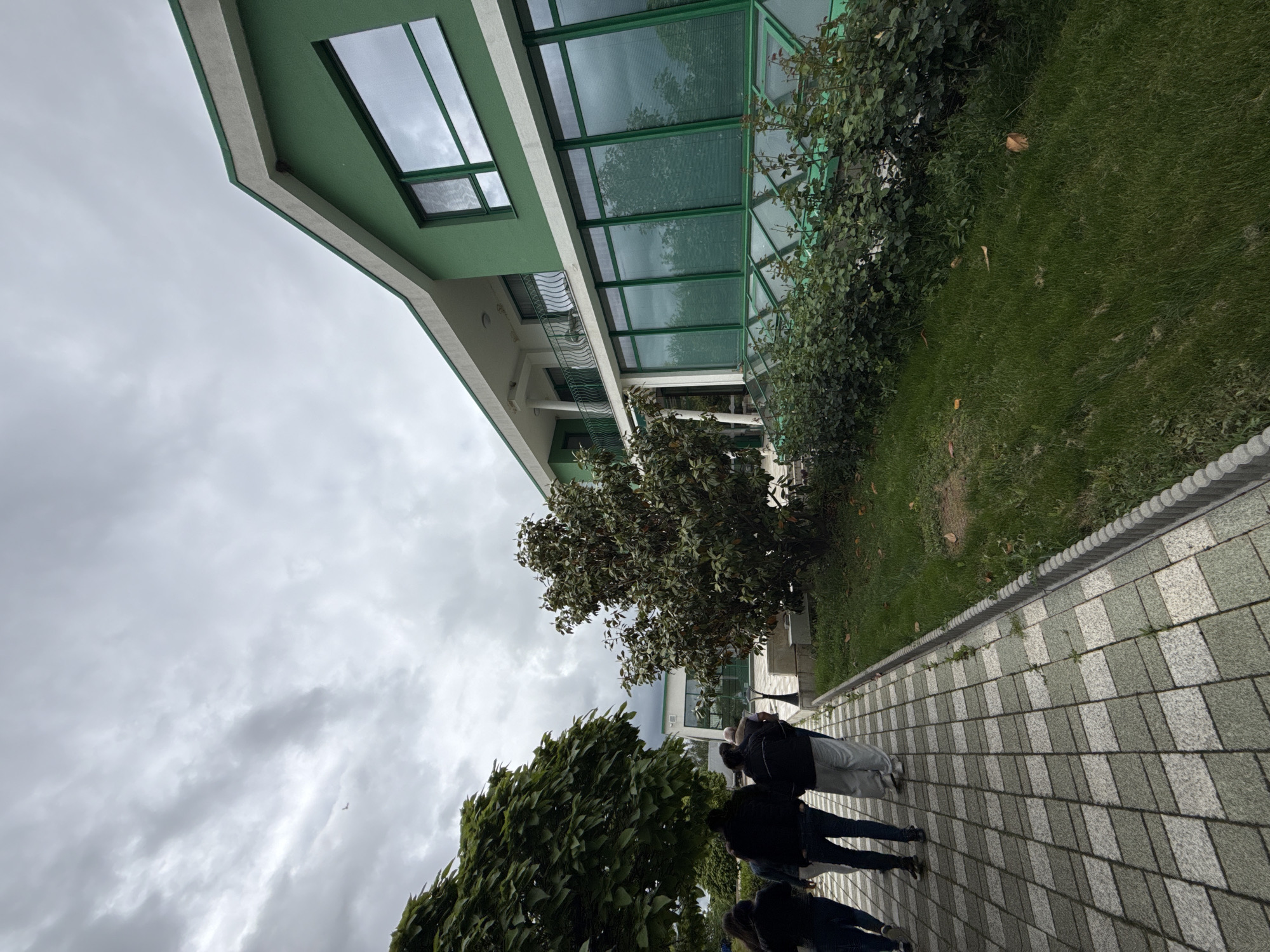 A low-angle shot shows the exterior of a building and a paved walkway, likely located in Gorna Mitropolia, Bulgaria. The building is green and white, with a modern design featuring large glass windows and a balcony with railings. A green roof extends over a covered area. Patches of grass and bushes are visible adjacent to the building. Four people walk along the paved walkway in the foreground. The walkway is constructed of light and dark grey square pavers. The sky is overcast with heavy clouds. Two trees flank the walking path, one on each side.
