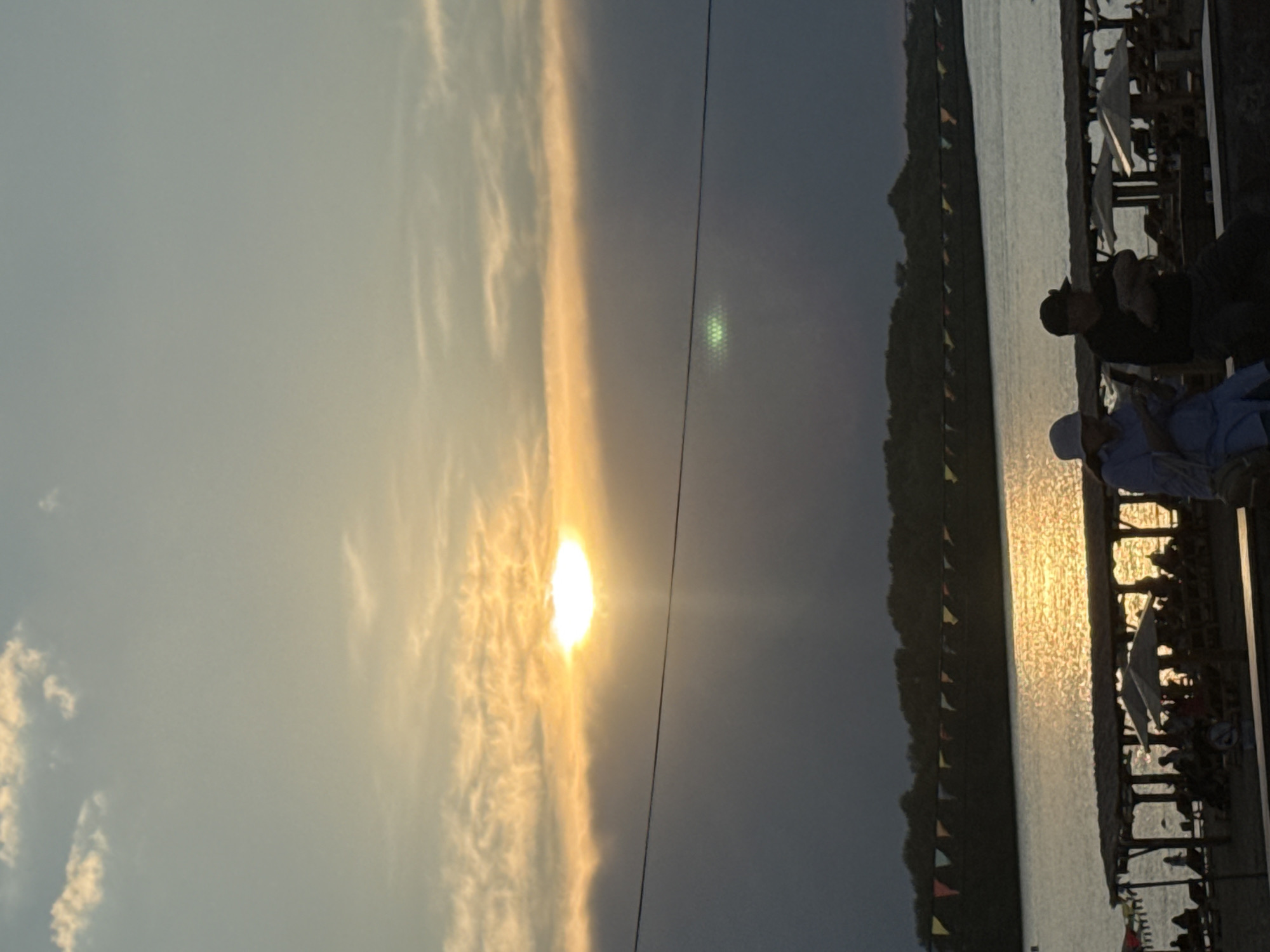 The image depicts a sunset scene by a body of water, likely a river or lake, in Pavlodar, Kazakhstan. The sun is low on the horizon, casting a warm, golden glow across the sky and reflecting on the water's surface, suggesting it's either early morning or late evening. Wispy clouds are illuminated by the sun, creating a dramatic effect.
On the right side of the frame, a wooden structure, possibly a pier or a boardwalk, extends over the water. Several people are visible on this structure, some seated and others standing. They appear to be relaxing and enjoying the sunset. The mood seems peaceful and serene.
Along the left bank of the water, a tree-covered hillside is visible, dotted with small, colorful triangular flags strung along its edge, adding a festive touch to the landscape. The water itself is calm, with the shimmering reflections of the setting sun creating a textured surface.
The overall scene conveys a sense of tranquility and natural beauty, with the vibrant colors of the sunset dominating the sky. The presence of people suggests a public or recreational area where locals gather to enjoy the evening. There is no visible text in the image.