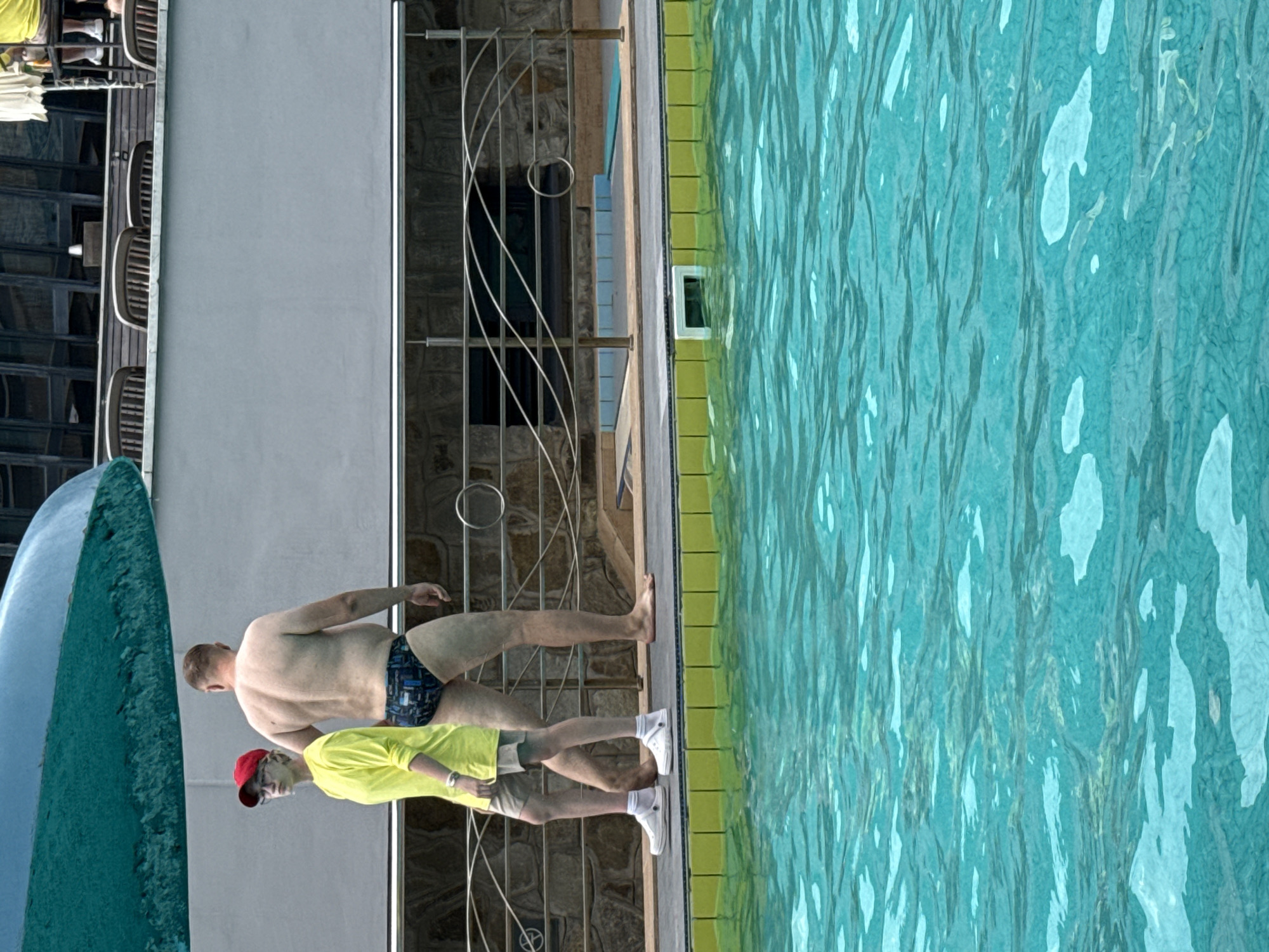 Two people are standing on the edge of a swimming pool. The person on the left is wearing a bright yellow long-sleeved shirt, tan shorts, white socks, and white shoes. This person also wears glasses and a red baseball cap. The person on the right is wearing black and blue patterned swim trunks and is bare-chested. They are both facing away from the camera, looking towards the pool. The swimming pool is filled with turquoise water. In the background, there is a metal railing, a textured wall, and chairs. The scene appears to be outdoors, possibly at a resort or public pool. The location is identified as Массандровский поселковый совет, Ukraine.