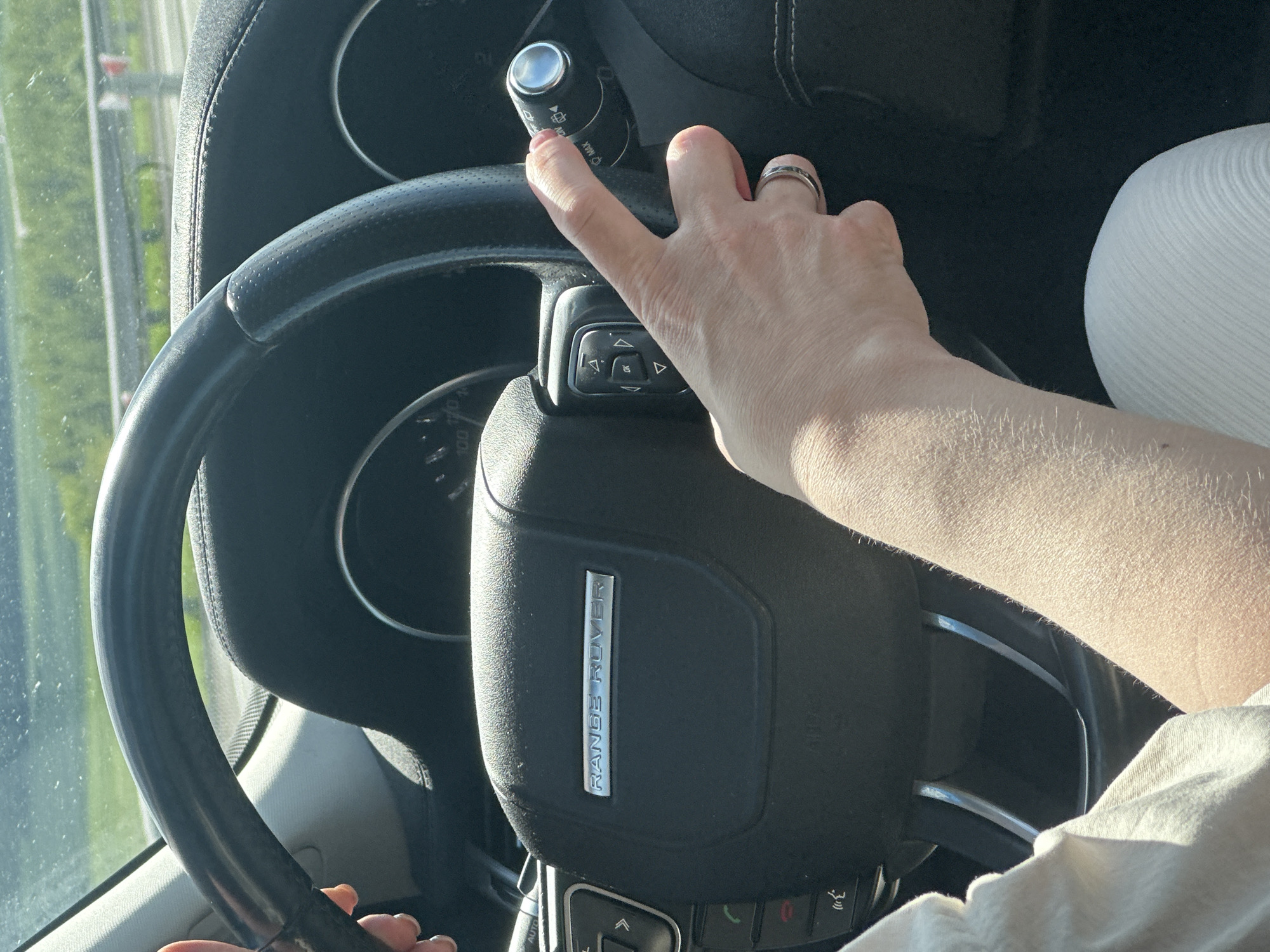The image shows the interior of a Range Rover vehicle. A person's hand is visible, manipulating the windshield wiper control stalk located to the left of the steering wheel. The steering wheel has the "RANGE ROVER" emblem prominently displayed on the center hub. The car appears to be in motion, as suggested by the blurred scenery visible through the windshield. The location is Unknown, Unknown.
