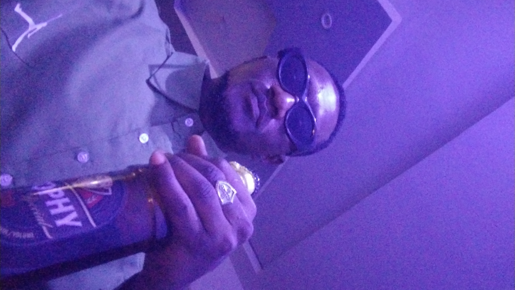 A dark-skinned man wearing sunglasses and a green collared shirt is holding a bottle of Trophy beer. He is wearing a ring on his right hand. The image is lit with purple light, possibly from a party or club setting. The man appears to be in the city of Jos, Nigeria. The background consists of a ceiling with recessed lighting.