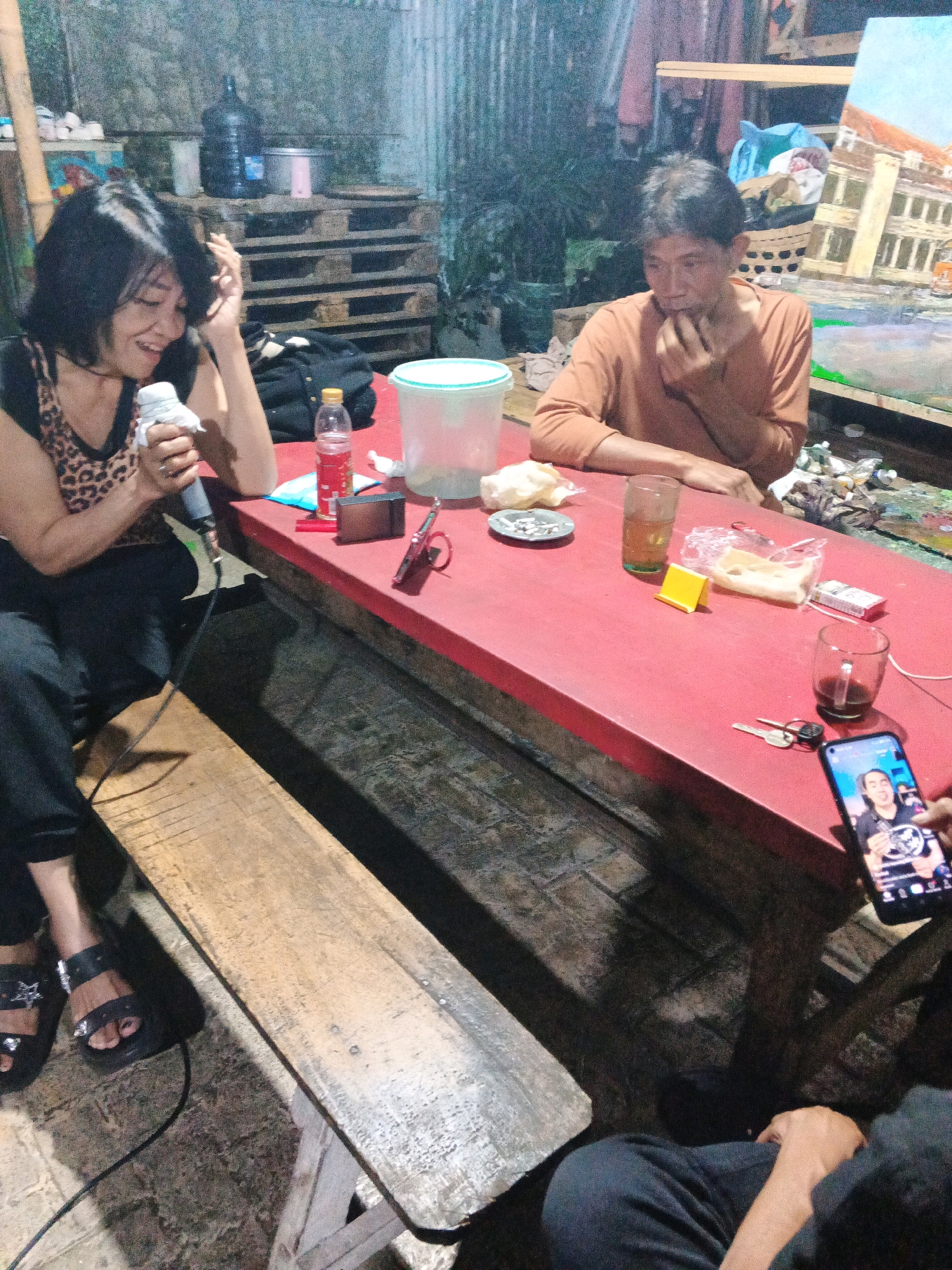In Ungaran, Indonesia, at night, two people sit at a long, red table. A woman with short black hair, wearing a leopard-print top and black pants, holds a microphone, smiling. A man with short dark hair, wearing an orange long-sleeved shirt, sits opposite her, looking down at his hands. On the table are various objects including a clear plastic container, a bottle of red liquid, a cigarette pack, glasses of liquid, keys, a smartphone displaying a video call with a person, and wrappers. The environment appears to be an informal outdoor or workshop setting, with wooden pallets stacked in the background and some artwork on display. The lighting is dim and artificial.