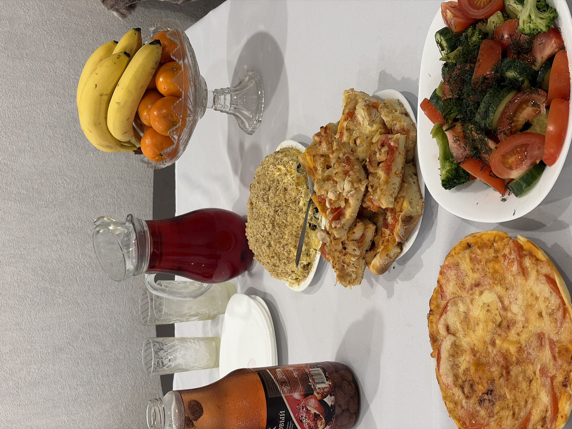 The image shows a table laden with food. In the upper left corner is a glass pedestal bowl filled with bananas and tangerines. To the right, a plate holds slices of a dish with bread, cheese, and tomato. Further right, there's a platter of salad containing broccoli, tomatoes, cucumbers, and carrots, seasoned with dill. In the lower right, a small pizza is visible. In the lower left, there's a pitcher of red juice, several empty glasses, and a stack of white plates. Next to it is a bottle of pomegranate juice. In the middle of the table is a cake covered in what appears to be grated nuts. The setting is likely a home in Sarapul, Russia, based on the bottle's labeling in Russian. There are no people visible. The scene presents a spread of food laid out on a white tablecloth, possibly for a meal or gathering.