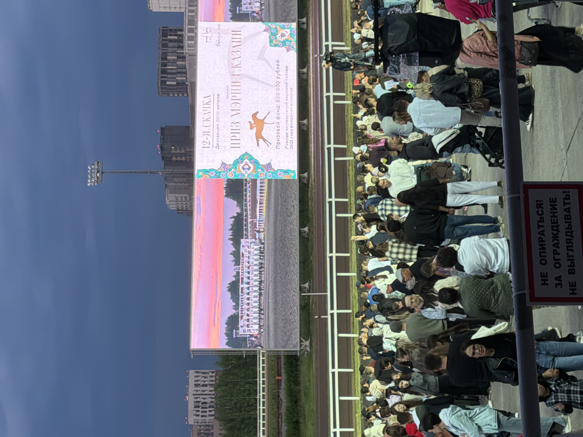 The image shows a crowd of people watching a large screen at a race track. The screen displays a horse race with jockeys and horses at the starting gate. Text on the screen indicates it is the "12th race" with a prize fund of 800,000 rubles. The sky is dark and cloudy, suggesting it is evening or a storm is approaching. The overall scene appears to be a public event with spectators gathered to watch the race on a screen, rather than the actual race.