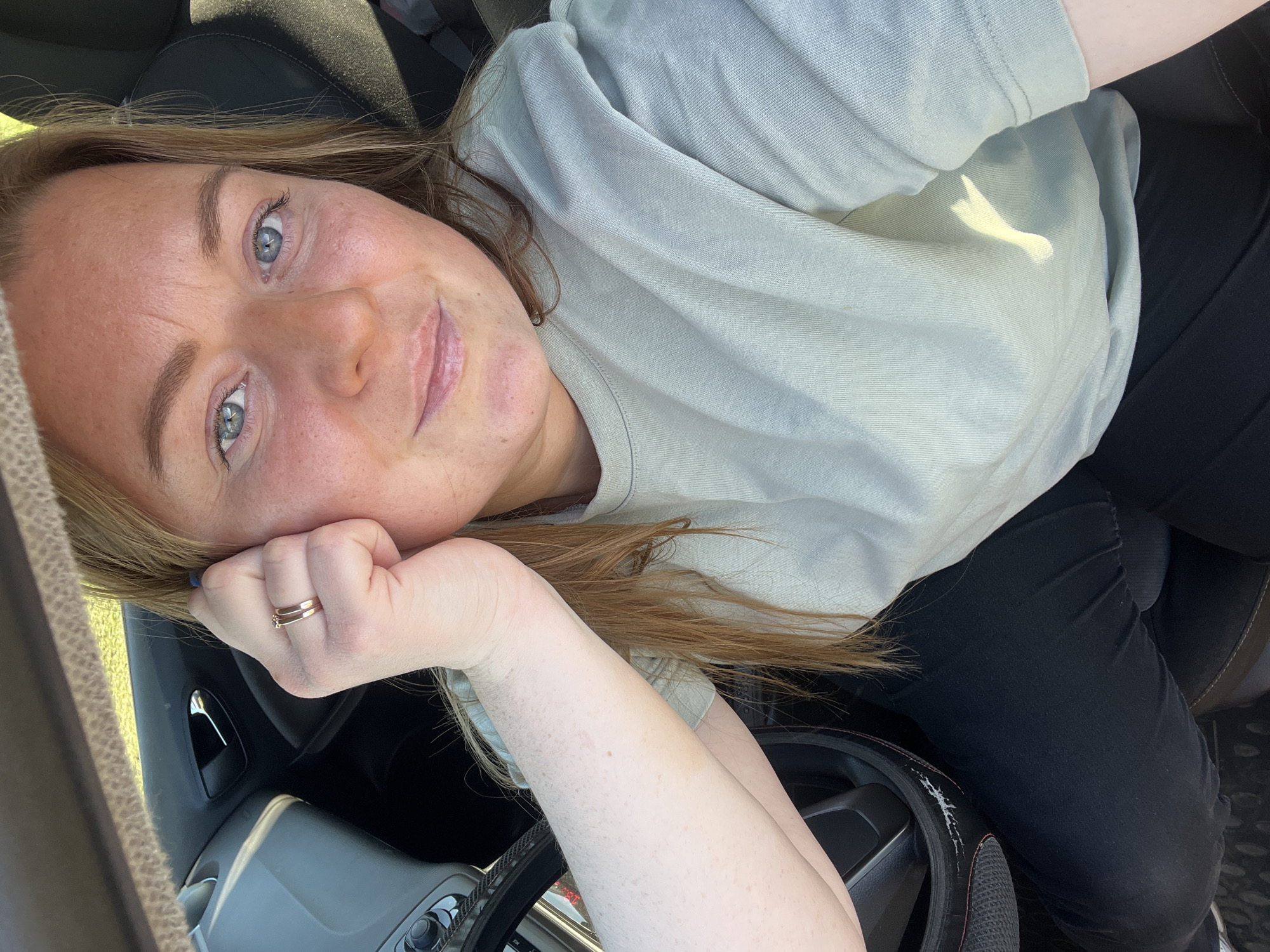 A light-skinned woman with blonde hair and blue eyes is reclining in the driver's seat of a car. She is wearing a light gray t-shirt and black leggings. Her left hand, adorned with a gold ring, is propping up her head. The interior of the car is visible, including the dashboard, steering wheel, and seat. The location is unknown and cannot be determined from the image.