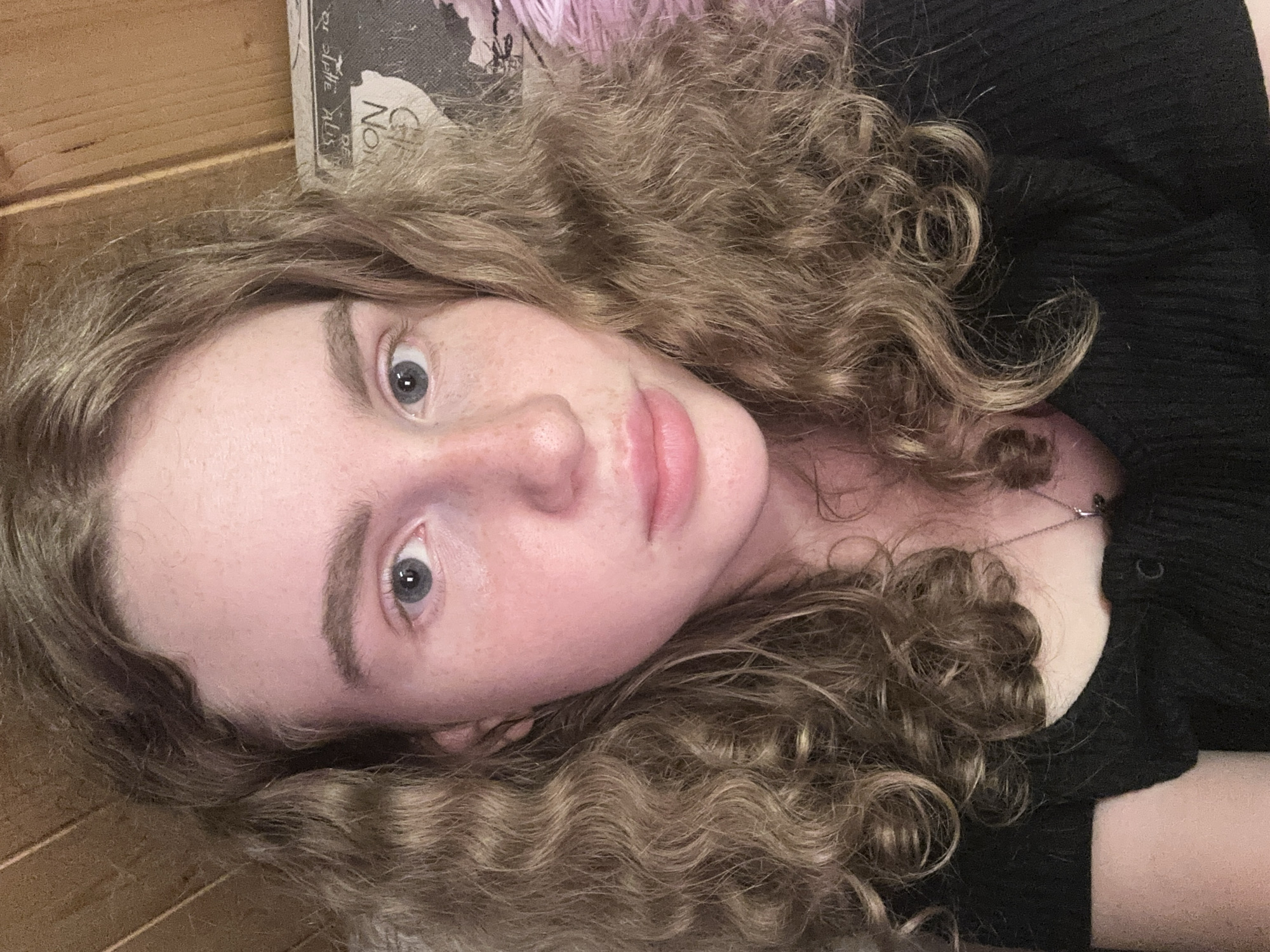 The image, rotated 90 degrees clockwise, presents a close-up portrait of a young person, likely female, with fair skin, scattered freckles across the nose and cheeks, and striking blue eyes. Her long, voluminous, light brown to blonde curly hair frames her face and spreads around her head. She is looking directly at the camera with a neutral and calm expression, her lips slightly parted.
She is wearing a dark, possibly black, ribbed top or sweater, and a delicate silver chain necklace with a small pendant is visible around her neck.
The background suggests an indoor setting. To the left (appearing at the top of the image due to rotation) is a wooden surface, possibly a wall or furniture. To the right (at the bottom of the image), a soft, dark fabric is visible, which could be a blanket or part of her clothing. Above her head, a book or framed item is partially visible, displaying faint text that includes "Noël" and possibly "lettres" or "slatte" along with "aus" or "rus". The lighting is soft and even, suggesting an indoor environment without strong direct light. No specific time of day or location cues are strongly evident beyond being indoors in what appears to be a residential setting, potentially Vasilyevo, Russia as per the context.
