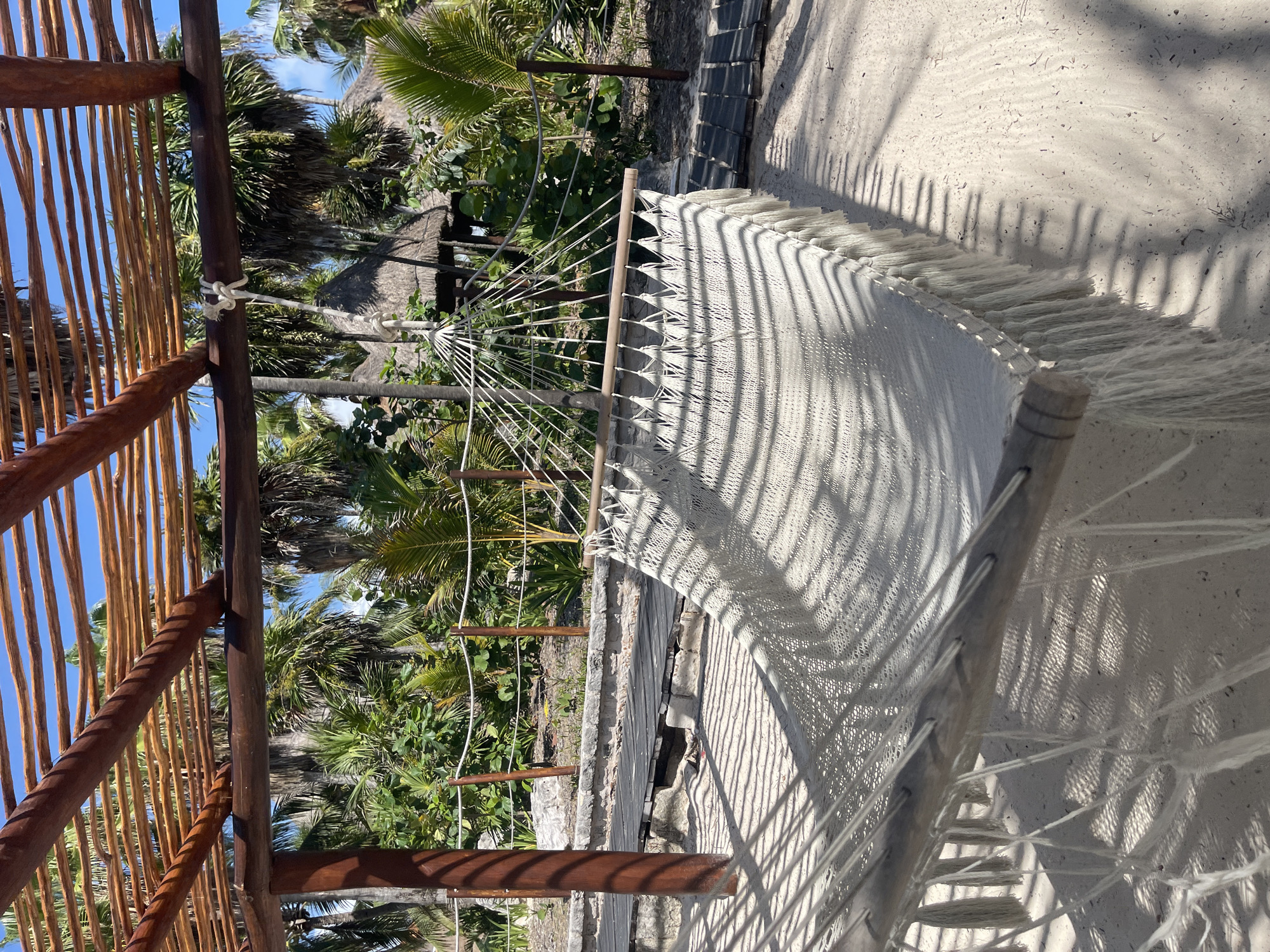 A hammock hangs under a wooden structure in Playa del Carmen, Mexico. The hammock is made of light-colored woven material and has fringe along the edges. The wooden structure is comprised of vertical and horizontal beams, some of which are made of thinner branches or sticks. The hammock is tied to the structure with white rope. Palm trees and other tropical vegetation are visible in the background. The ground is sandy and covered in shadows cast by the surrounding foliage.
