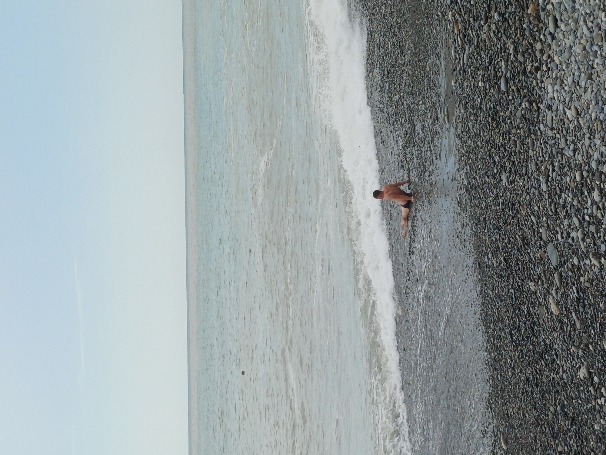 A man is sitting on a pebble beach with waves washing over his legs. The sky is clear and the ocean is calm. The scene appears to be a beach in Sochi, Russia. The man is wearing black swim trunks. There are no other people or objects visible in the image. The time of day is unclear but appears to be daytime.