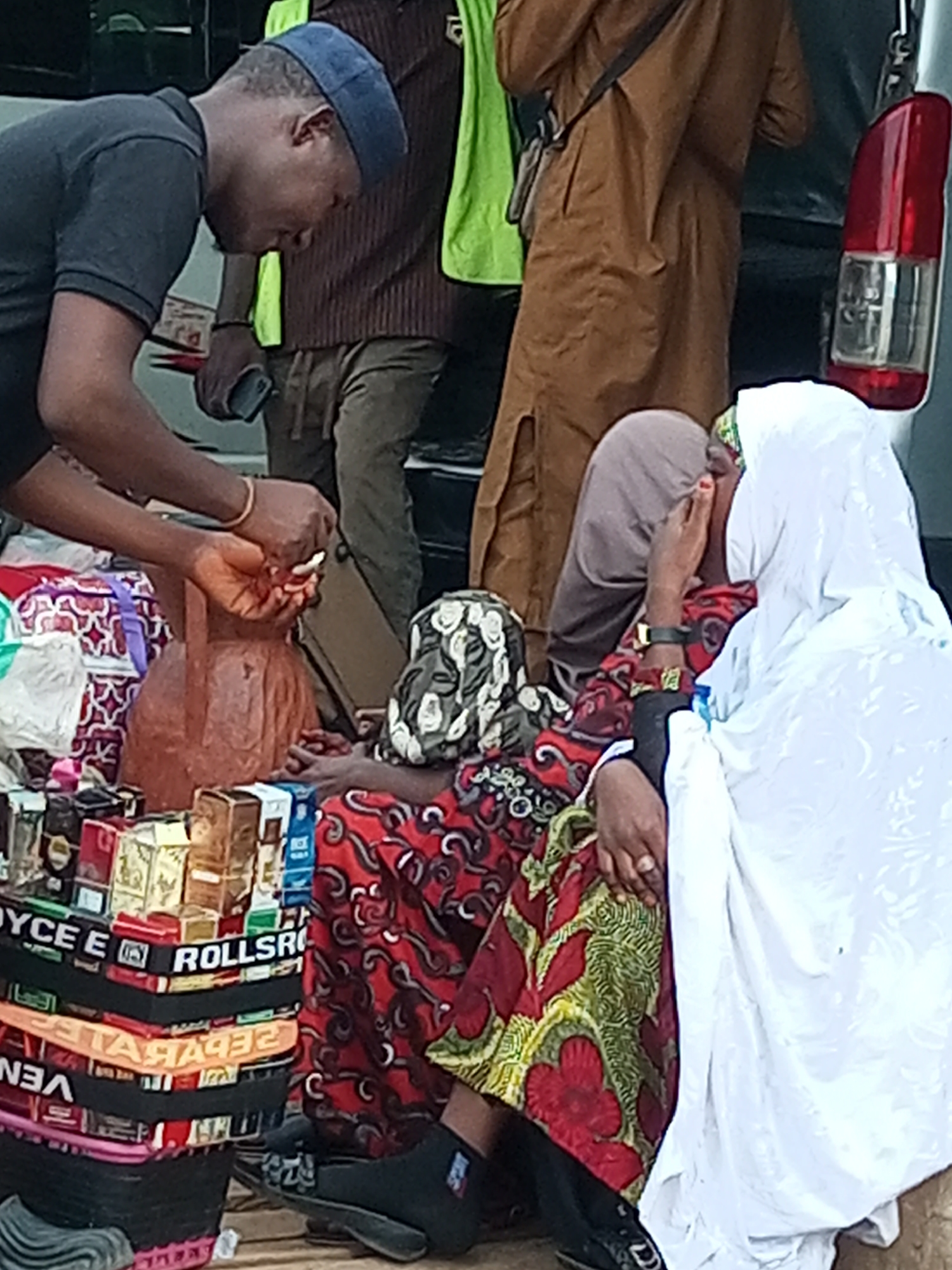 In Gombe, Nigeria, a man wearing a dark blue cap and a black t-shirt is leaning over a stall. He is holding a small object, possibly a phone, in his right hand, and his left hand is extended towards a clay pot. Behind him, a person in a brown tunic and another in a bright green vest are visible. In front of the man, at the stall, two women are seated. One woman, wearing a patterned headscarf and a red and green floral dress, is covering her face with her hand. The other woman, to her right, is almost entirely covered by a white hijab, with only a portion of her face visible. The stall displays various items, including stacks of cigarette packs and what appear to be colorful fabric trims. The scene suggests a market or street-side transaction.