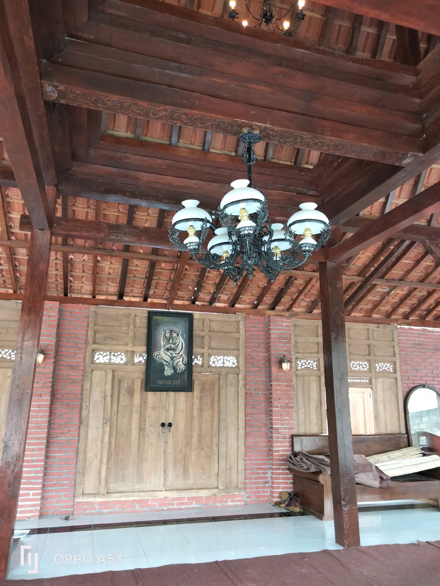 The image depicts the interior of a structure with a high, wooden-beamed ceiling covered with red roof tiles. A large, ornate, black chandelier with multiple white lamps hangs from the ceiling. The walls are constructed from red brick and feature large wooden doors with decorative panels. A framed painting of a tiger with the word "INDONESIA" below it hangs on one of the wooden walls. To the right, a wooden bench with folded blankets and some rolled-up mats is visible. A mirror is propped against the wall behind the bench. The floor is tiled with light-colored square tiles. The scene suggests an outdoor or semi-outdoor covered area, possibly a traditional Indonesian building. The location in Trenggalek, Indonesia, is not directly indicated by any visual cues in the image itself, but it is provided as context for the scene.