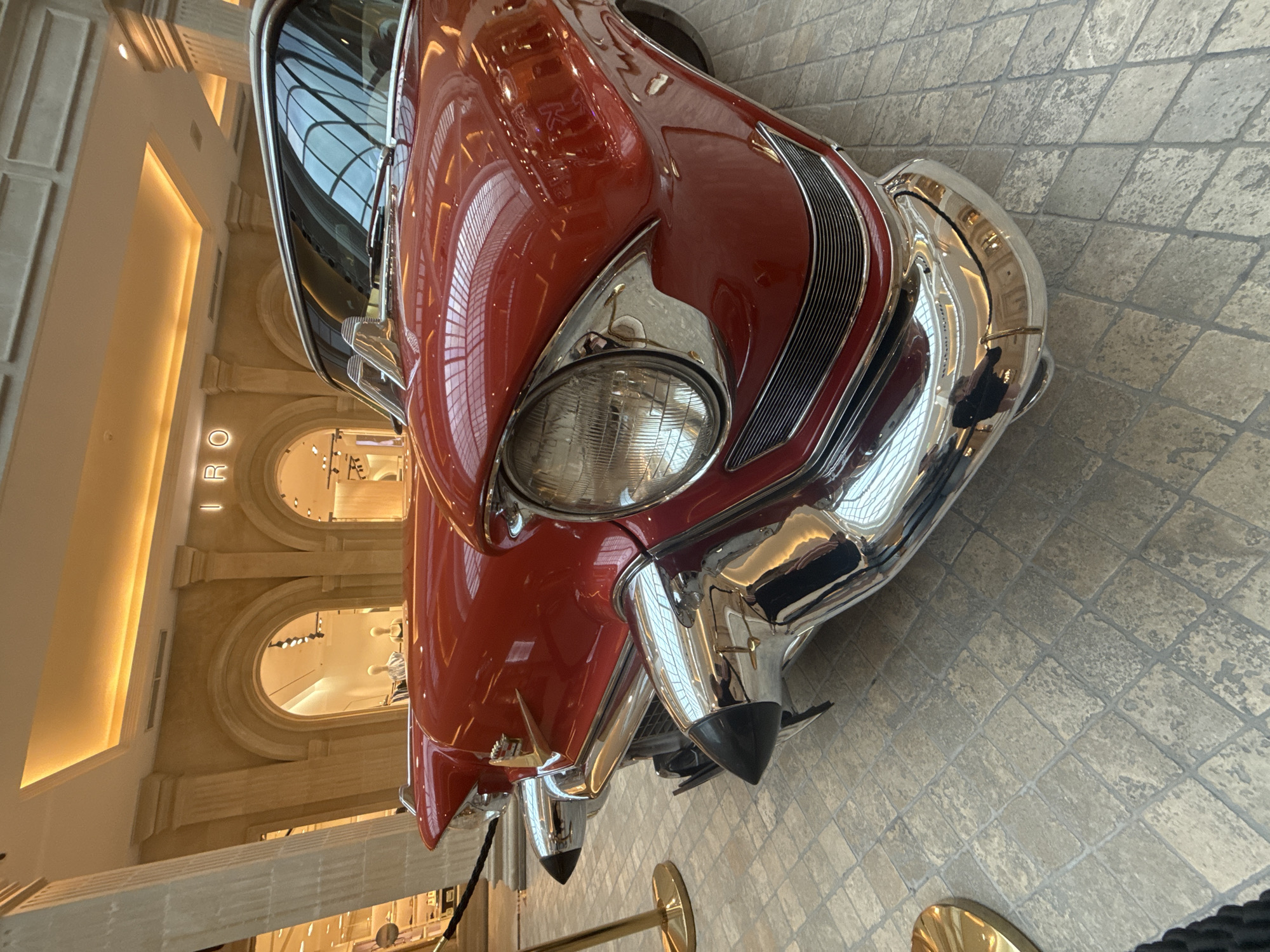 A close-up, low-angle shot shows the front of a vintage, bright red car, its chrome gleaming. The car's hood is angled towards the camera, and the prominent headlight and curved grille are in focus. The car is parked on a floor tiled with large, square, grey stones. In the background, a building interior is visible, featuring classical architectural elements like arches and columns. Several arched windows reveal illuminated retail displays with mannequins. The overall lighting suggests it is daytime, with artificial lights in the building interior casting a warm glow. The scene appears to be indoors, possibly in a museum, gallery, or a high-end shopping center. There is no visible text in the image.