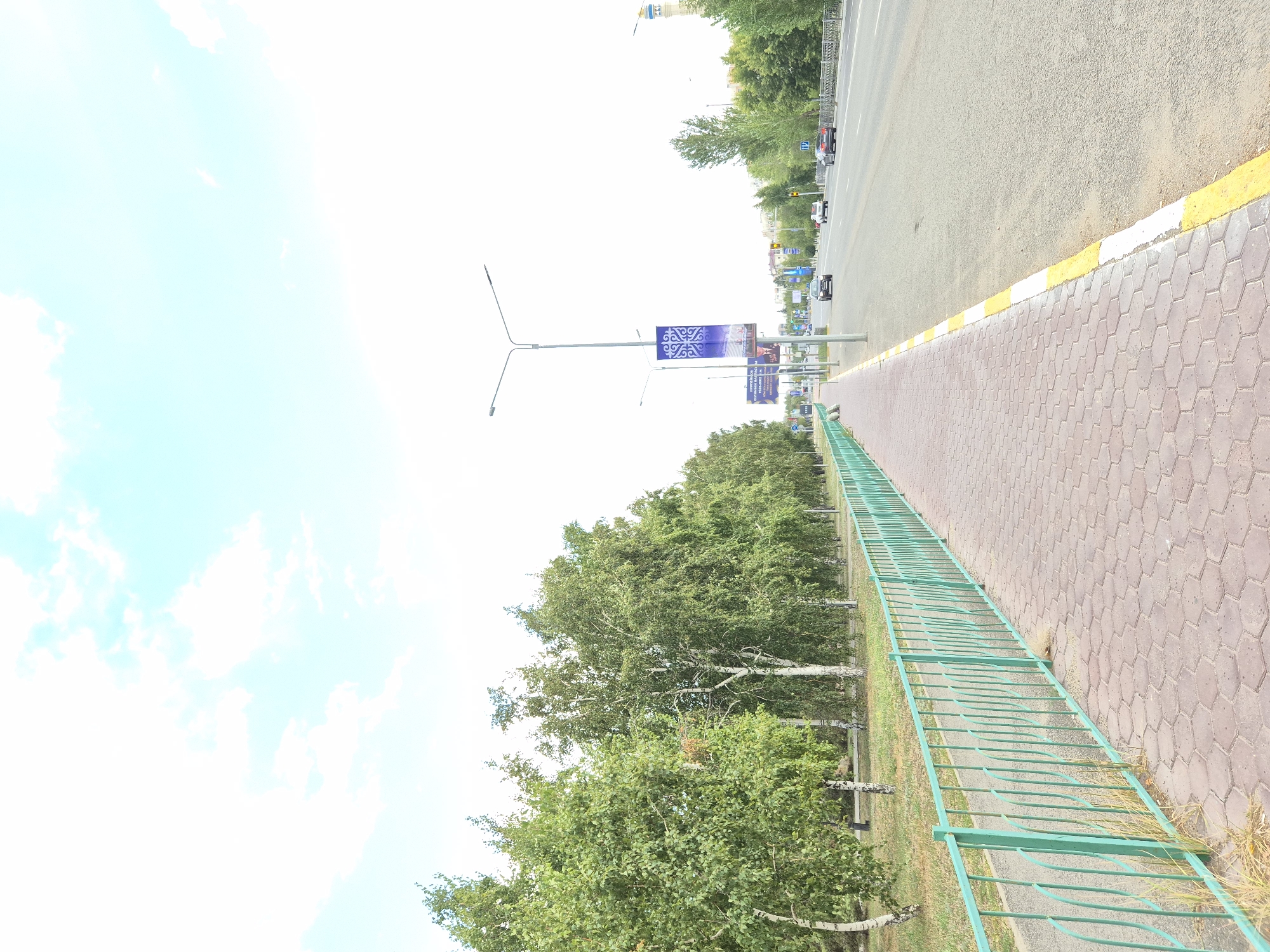 The image captures a street scene in Kokshetau, Kazakhstan. On the left, a paved sidewalk made of hexagonal brick pavers runs alongside a green metal fence. Beyond the fence, a row of birch trees with green foliage lines the path. The sidewalk transitions to a road with a yellow and white painted curb. On the road, several cars are visible, including a black sedan, a silver sedan, and a dark-colored SUV. Streetlights and decorative banners, one featuring a blue and white traditional pattern, are placed along the roadside. In the distance, more trees and buildings can be seen. The sky is bright with scattered white clouds.