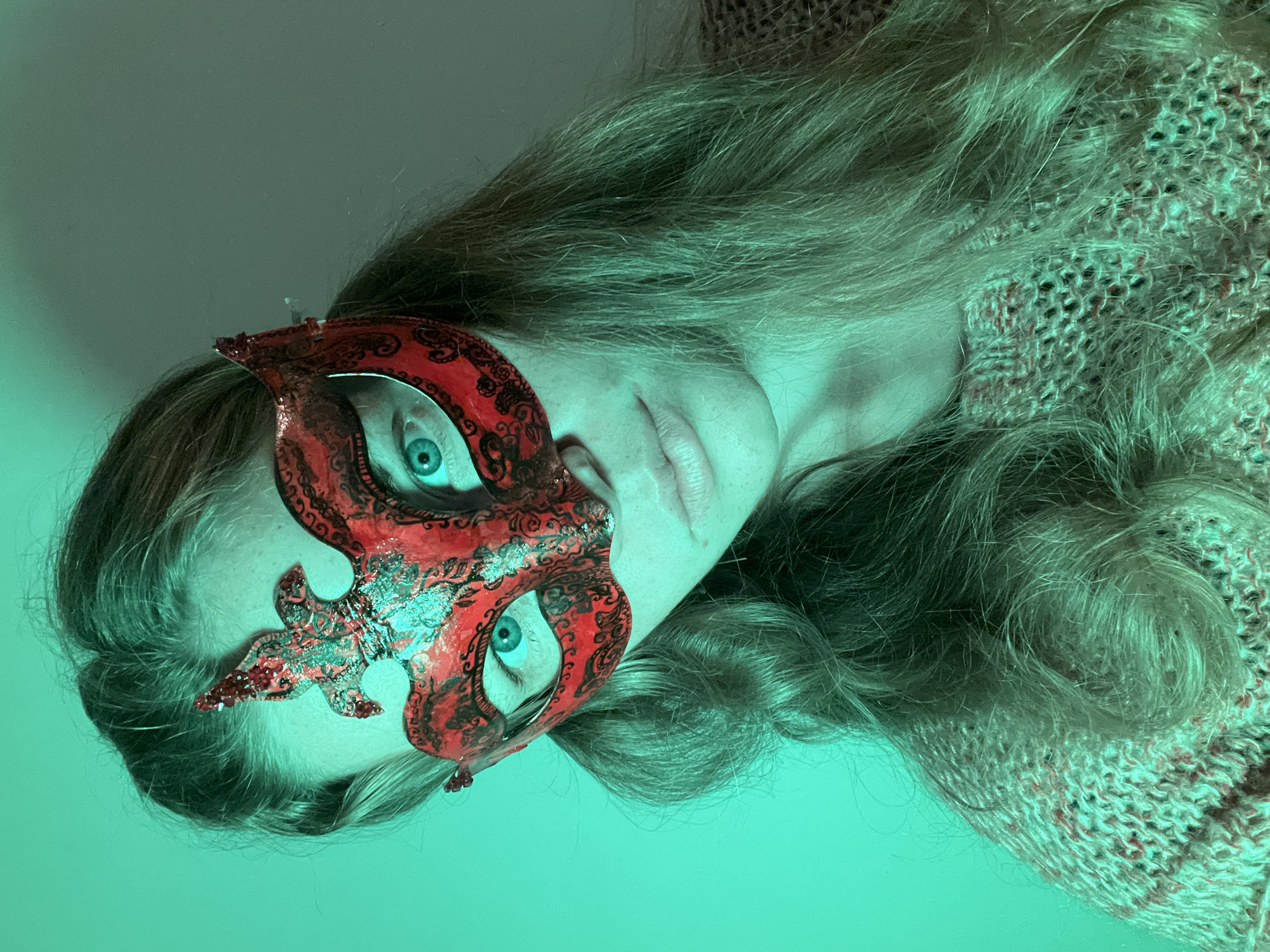 A person wearing a red and black ornamental mask is visible in a close-up shot. The mask covers the upper left portion of their face, leaving one eye exposed. The person has fair skin and blonde hair. They are wearing a knitted garment. The lighting is green, creating an eerie effect. The location is not possible to identify as Budva, Montenegro, from this image alone.