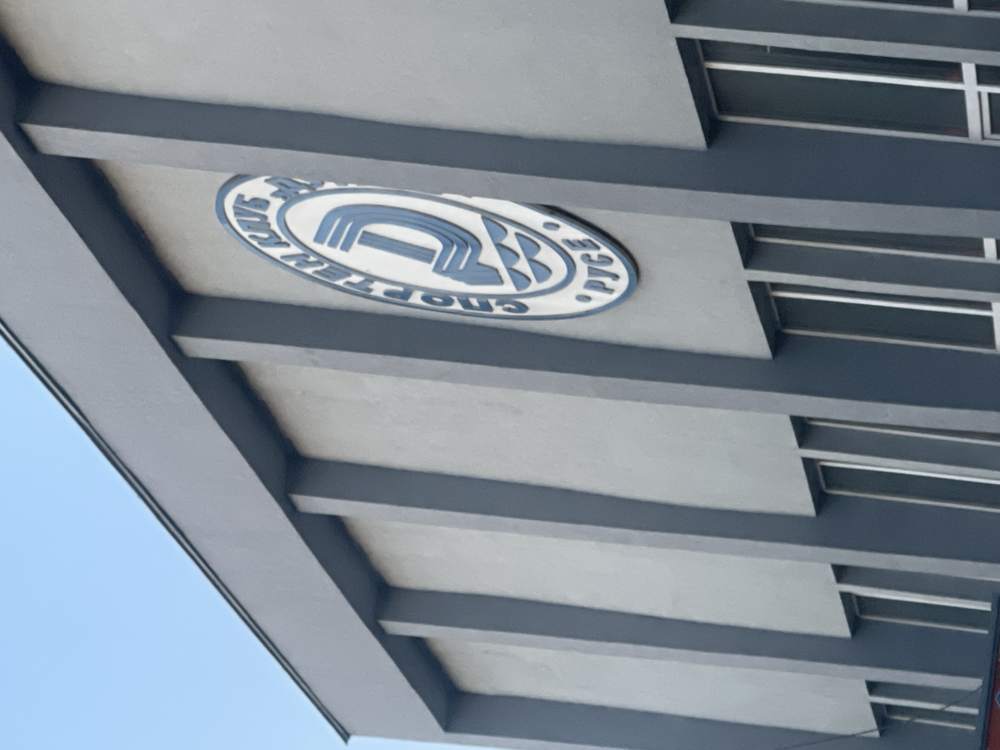 The image shows the facade of a building with a prominent blue and white circular emblem. The emblem features the Cyrillic letters "СПОРТЕН КЛУБ" (Sport Club) in a circle around a stylized letter "Д" and the word "РУСЕ" (Ruse) at the bottom. The building itself is grey with dark blue vertical accents and several large rectangular windows. The sky is clear and blue. The emblem is likely on the building of a sports club in the city of Ruse, Bulgaria. There are no people visible in the image.