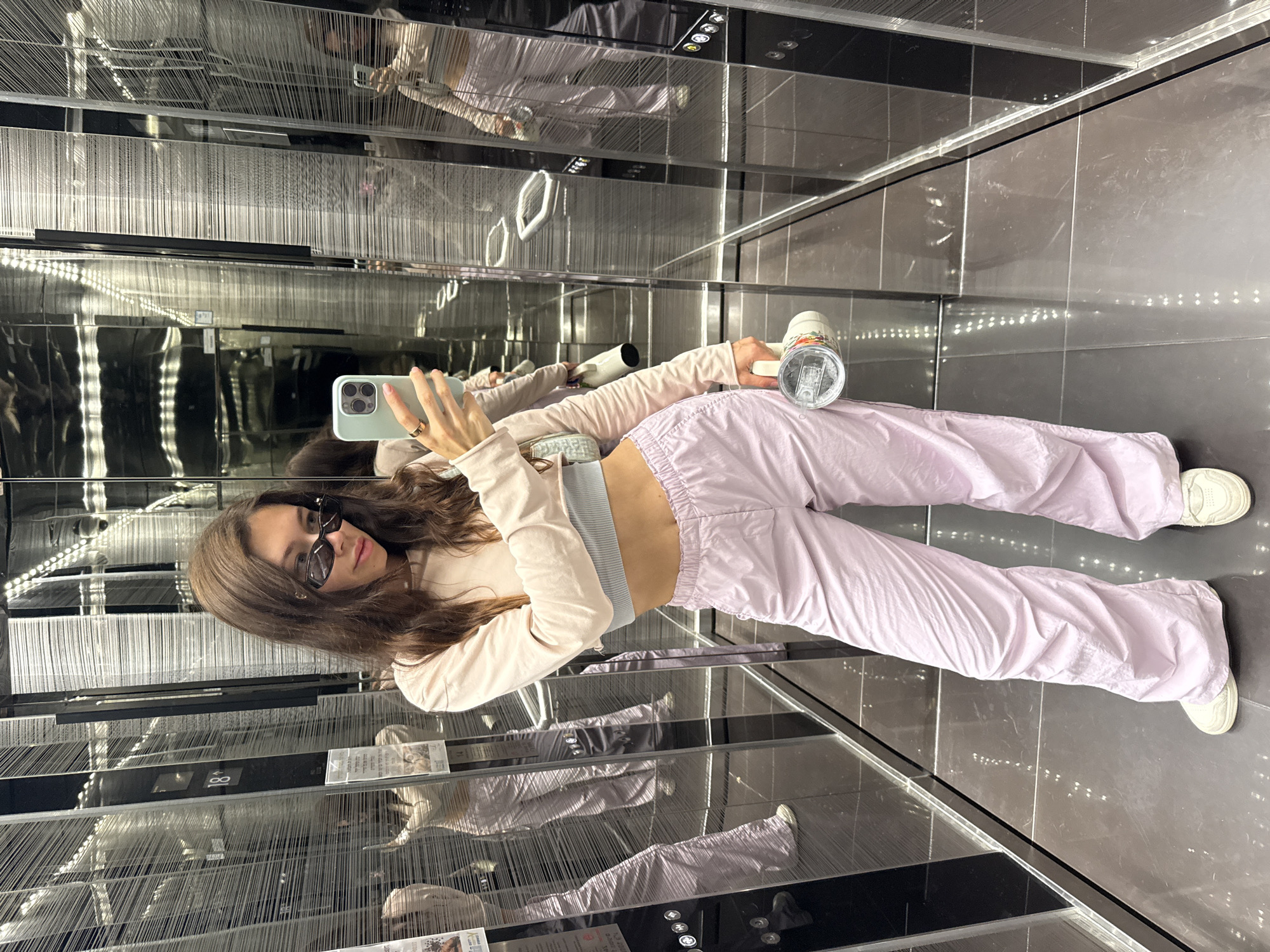A woman wearing sunglasses, a pink cropped sweater, a grey tank top, and pink pants is taking a selfie in an elevator. She is holding a light green smartphone in her left hand and a white water bottle in her right. The elevator walls are reflective, creating multiple reflections of the woman and the environment. The elevator interior is modern with black tiled floors and decorative vertical strands on the walls. The elevator display on the wall shows the number 18. From researching the image, the location is the Brown Hotel in Tel Aviv, Israel.