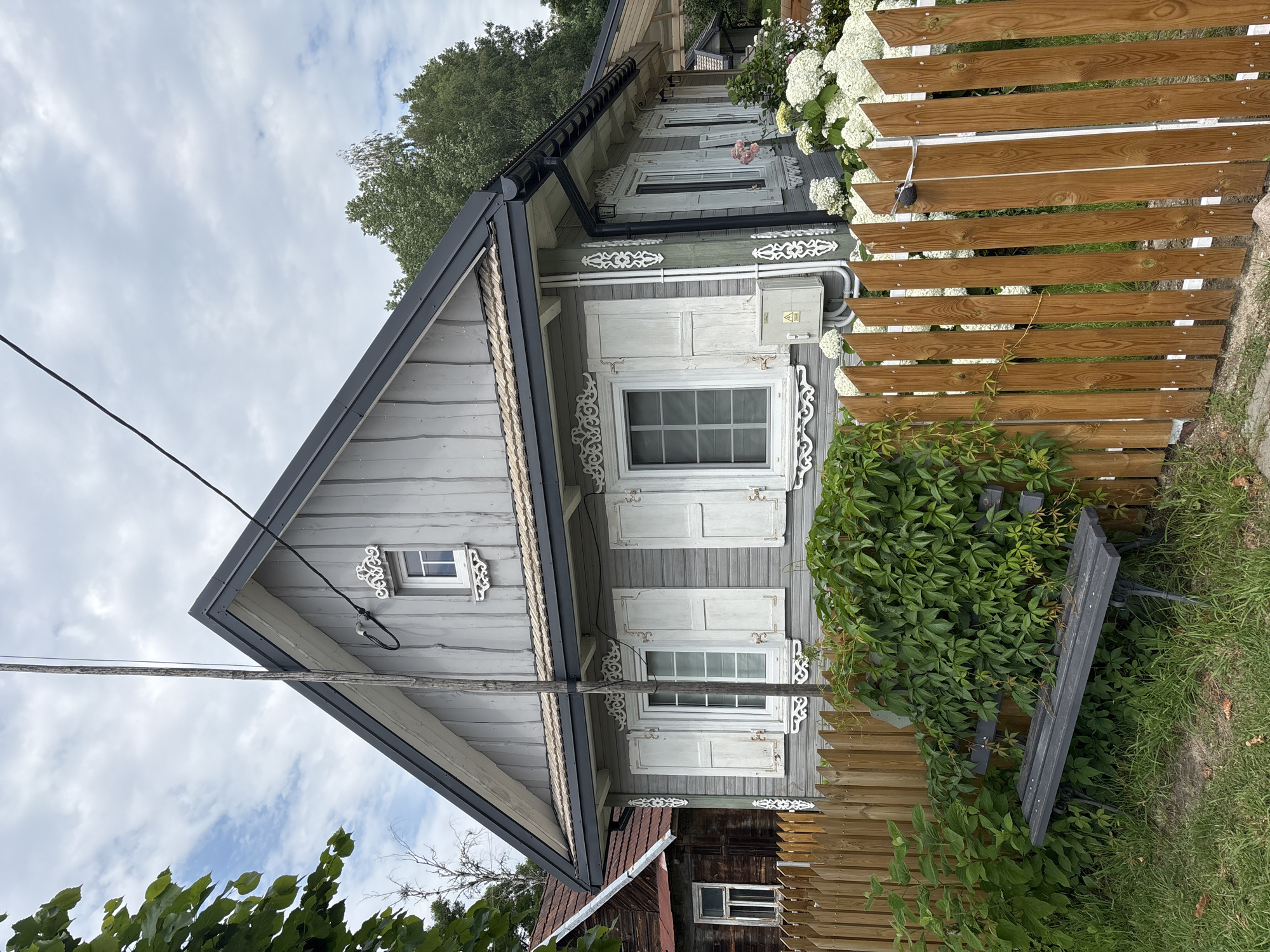 The image depicts a traditional wooden house in Trześcianka, Poland. The house is painted gray with white shutters and decorative trim around the windows. A wooden fence encloses the property, with a lush green vine growing over a dark-colored bench placed near the fence. In the background, a neighboring wooden house with a red roof is visible, along with trees and a cloudy sky. The scene appears to be a quiet, rural setting.