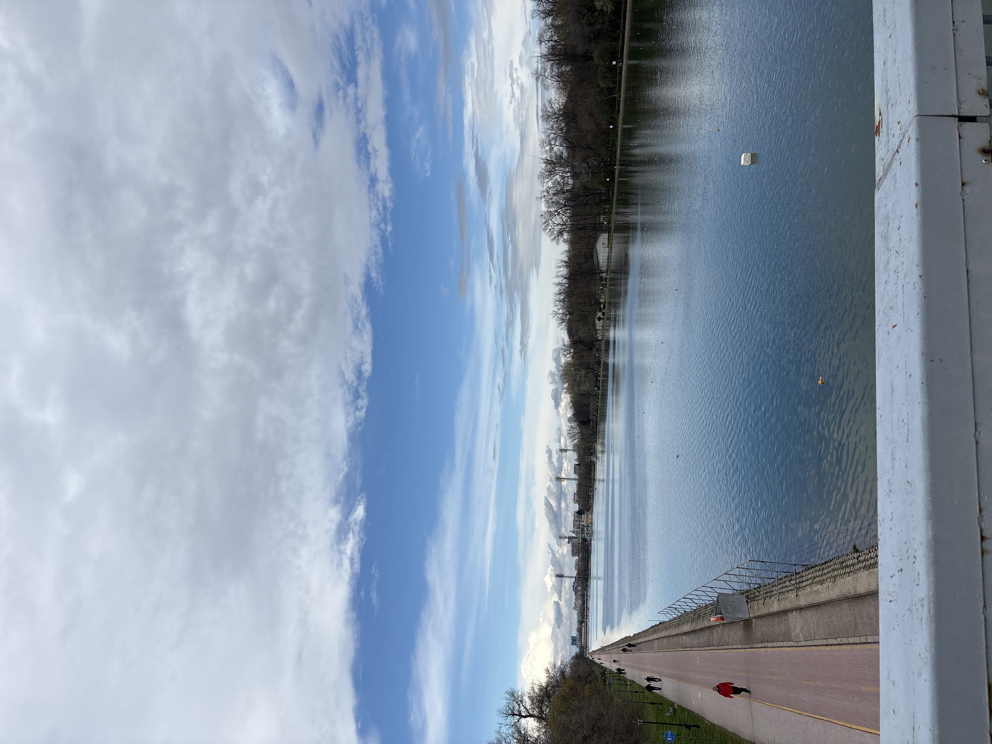 The image shows a view of the Rowing Canal in Plovdiv, Bulgaria, from an elevated perspective, possibly a bridge. The canal is a long, straight body of water with reflections of the sky and surrounding trees. Along one side of the canal is a walkway with several people walking or running. A person wearing a red jacket is visible on the walkway. The sky is partly cloudy, with patches of blue visible. The environment includes trees lining the canal, and the city skyline can be seen in the distance. A white object is floating on the water's surface.