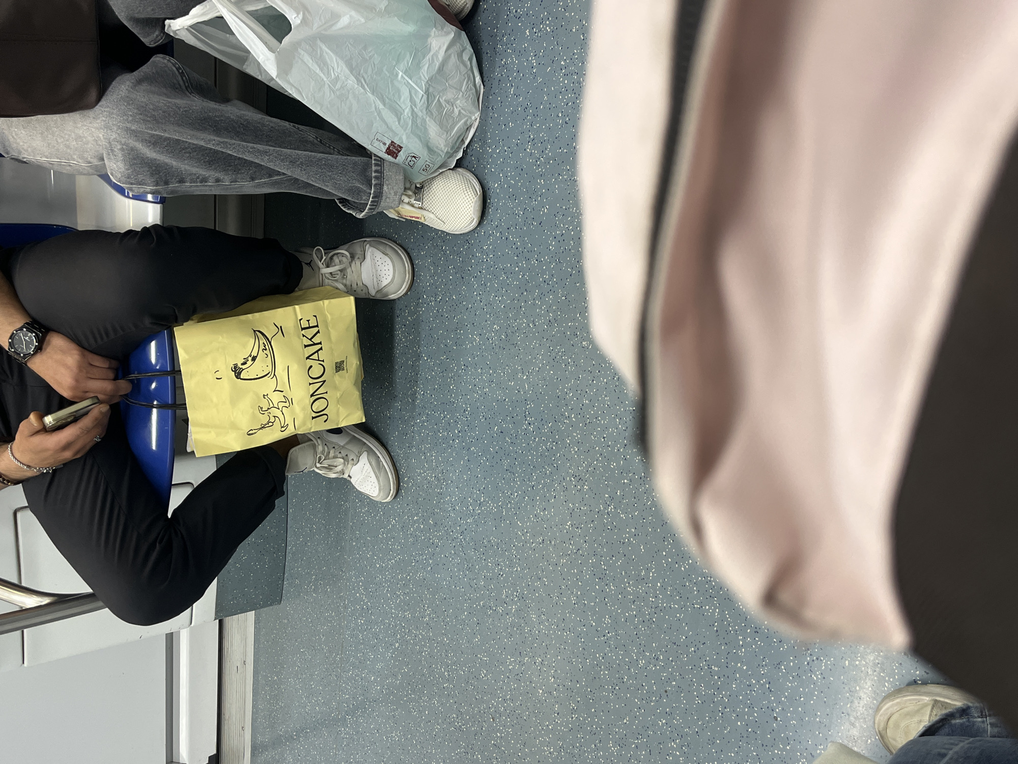 The image shows a section of the interior of a vehicle, possibly a subway or train. There are three people visible. One is sitting in a blue seat, wearing black pants and white sneakers and holding a phone. A yellow bag with the word "JONCAKE" printed on it is between their legs. Another person is visible sitting next to them wearing gray jeans and white sneakers, holding a white plastic bag. The third person, only partially visible on the right side, is wearing jeans and sneakers. The floor is a speckled grey-blue. Based on the Joncake bag, it is possible that the location is Barcelona, Spain, where Joncake bakeries exist.