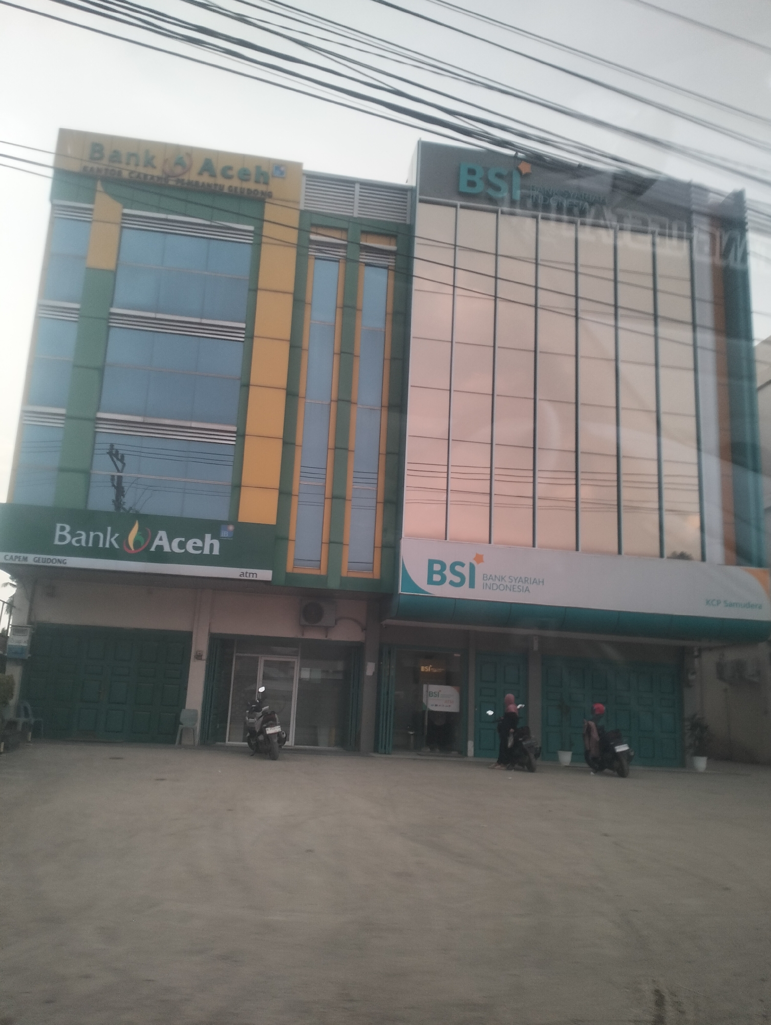 This media file shows the exterior of two adjacent bank buildings in Blangpeuria, Indonesia. The building on the left is the "Bank Aceh" and the building on the right is "BSI BANK SYARIAH INDONESIA". Both buildings have multiple stories with large windows. The Bank Aceh building has a green and yellow facade, while the BSI building has a modern glass facade. In front of the buildings, there are several parked motorcycles. The ground in front of the buildings appears to be a paved area. The sky is visible at the top of the image, with some electrical wires crossing the frame. The overall lighting suggests it is daytime. The scene depicts a typical street view of financial institutions in a commercial area. Visible text includes the names of the banks, "Bank Aceh", "BSI", "BANK SYARIAH INDONESIA", and the branch information "KANTOR CABANG PEMBANTU GEUDONG", "CAPEM GEUDONG", and "KCP Samudera". There is also "atm" visible below the Bank Aceh signage.