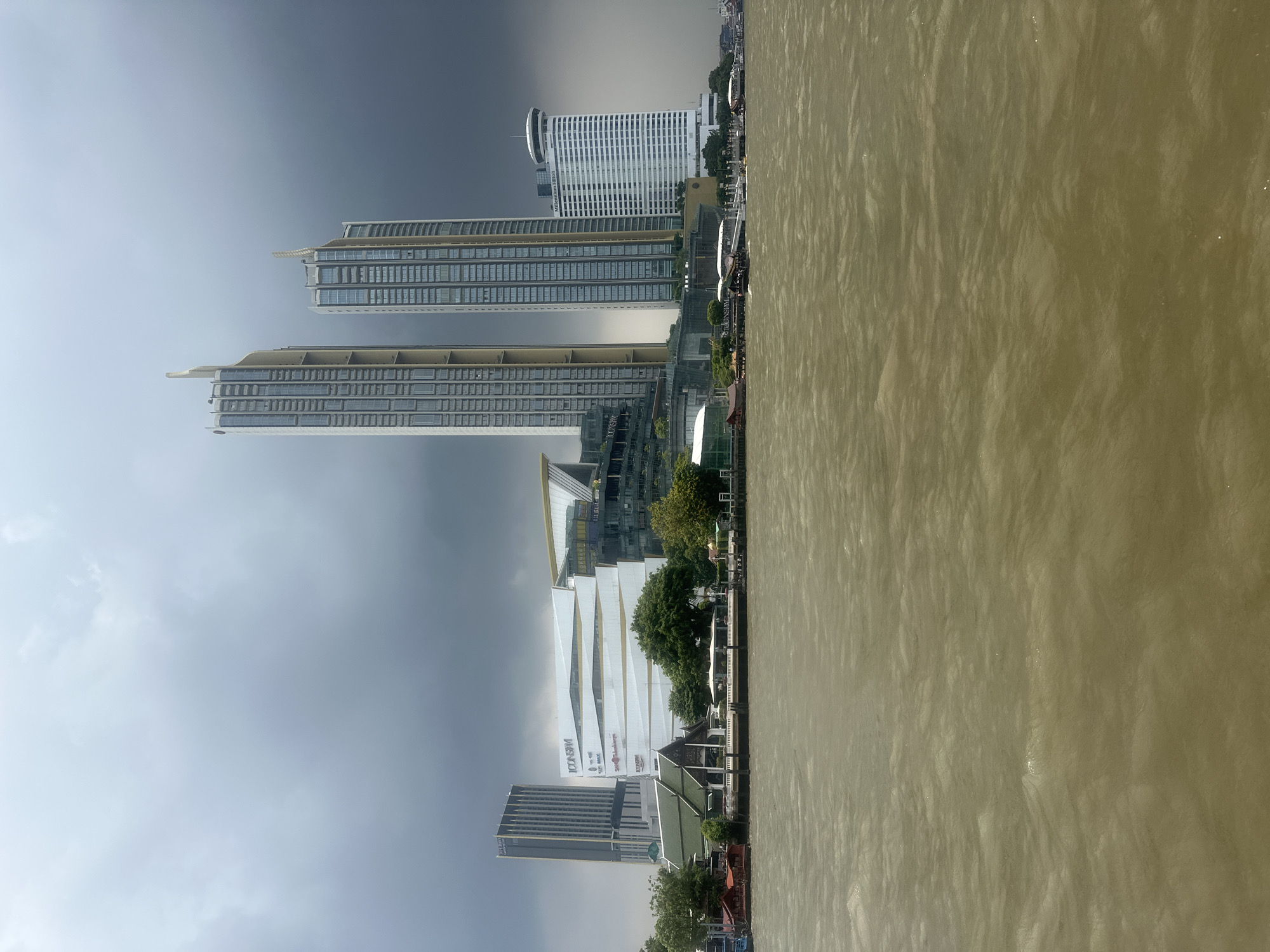 This image presents a panoramic view of a modern cityscape from across a wide river, under an overcast sky. On the far bank, a series of prominent buildings dominate the horizon.
Several tall, contemporary skyscrapers with numerous reflective windows rise vertically, showcasing modern architectural styles. Among these, a large, distinctive white building with a wavy, tiered facade is clearly visible, featuring the prominent signs "ICONSIAM" and "Siam Takashimaya." Further along the riverbank, another white high-rise building with a unique circular structure at its top can be seen, bearing the text "MILLENNIUM HILTON." Interspersed with these modern structures are areas of green foliage and trees, as well as some buildings featuring traditional Thai-style roofs closer to the river's edge.
The foreground is dominated by the river, which appears wide and has a murky, brownish-green color, with a textured, slightly choppy surface. The sky above is heavily clouded, varying from a lighter grey on the left to a darker, more dramatic grey on the right, indicating an overcast or potentially stormy day. The scene is captured during daylight hours.
The visible landmarks, particularly "ICONSIAM" and "MILLENNIUM HILTON," strongly suggest the location is along the Chao Phraya River in Bangkok, Thailand. No specific human activity or people are discernible in the static scene.