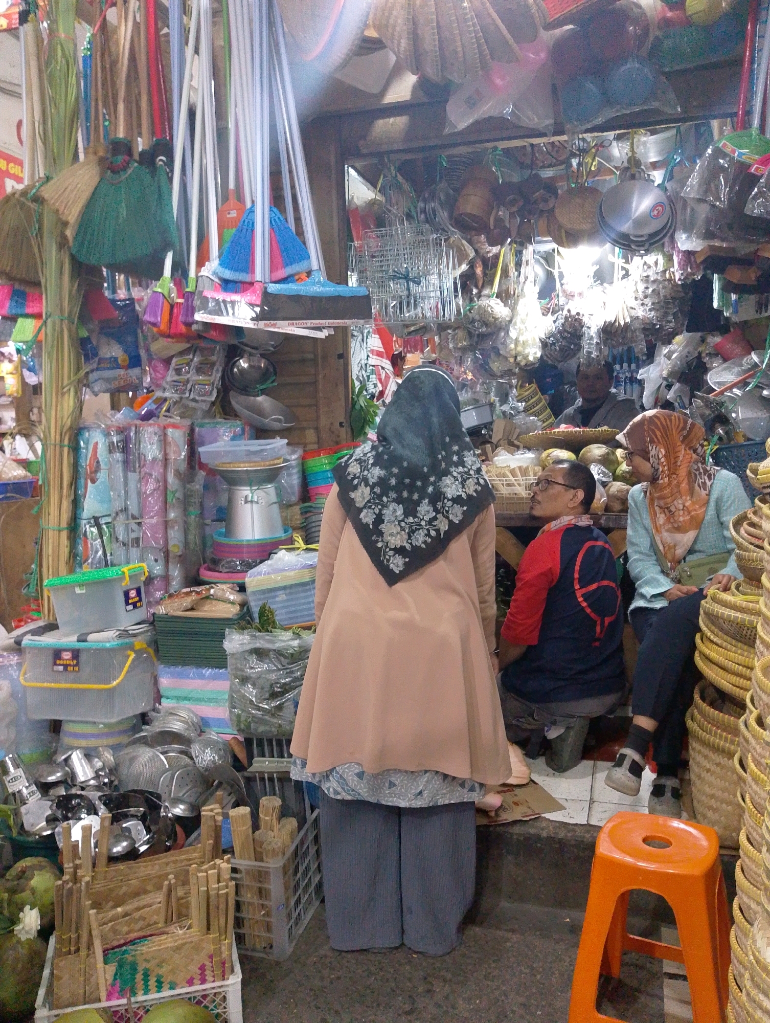 This image depicts a bustling street-side market stall in Lembang, Indonesia, specializing in household goods and kitchenware. A woman wearing a light brown abaya and a black floral hijab stands with her back to the camera, facing a stall filled with various items. To her right, a woman in an orange hijab is seated, conversing with a man wearing a red and blue shirt and glasses who is also seated. The stall is densely packed with merchandise, including stacks of plastic containers, woven baskets, metal pots and pans, brooms, and what appear to be food items like coconuts and bundles of dried sticks. The environment suggests a humid, outdoor market setting with a variety of textures and colors from the stacked goods.
