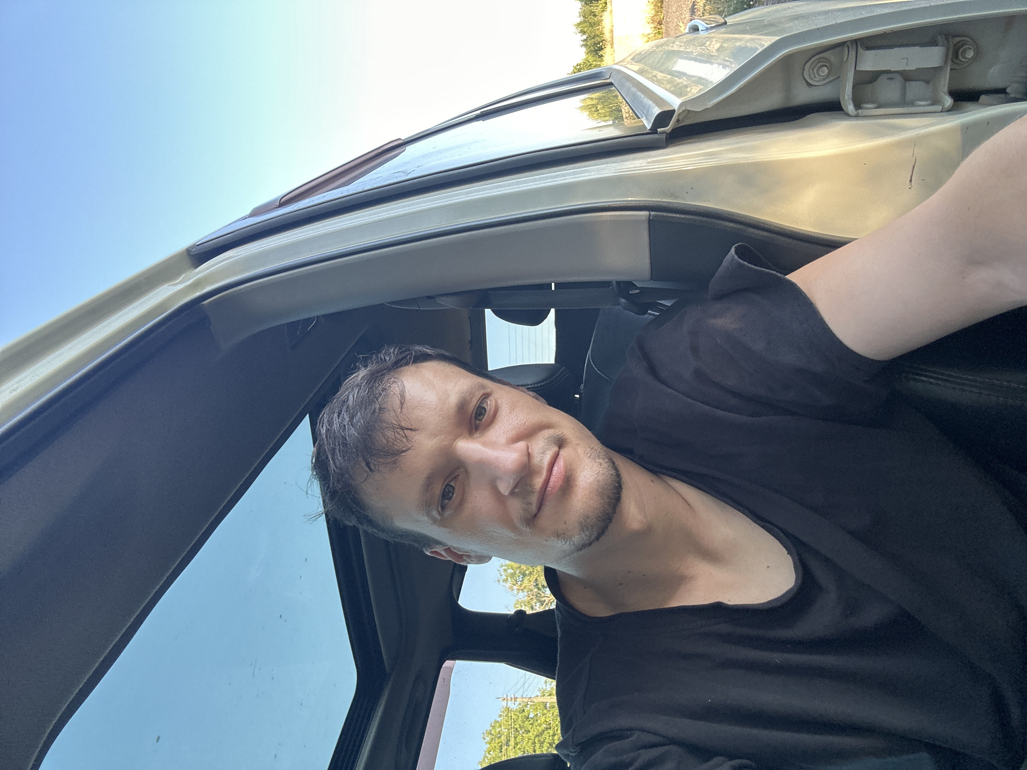 A man with short, dark hair and stubble is in the driver's seat of a car, looking at the camera with a slight smile. He is wearing a black t-shirt. The car has a sunroof, which is open, and the passenger door is ajar. The background outside the car appears to be rural, with trees and a bright blue sky. The image appears to have been taken on a sunny day. The location of Makiv, Ukraine is not visually discernible from the image.
