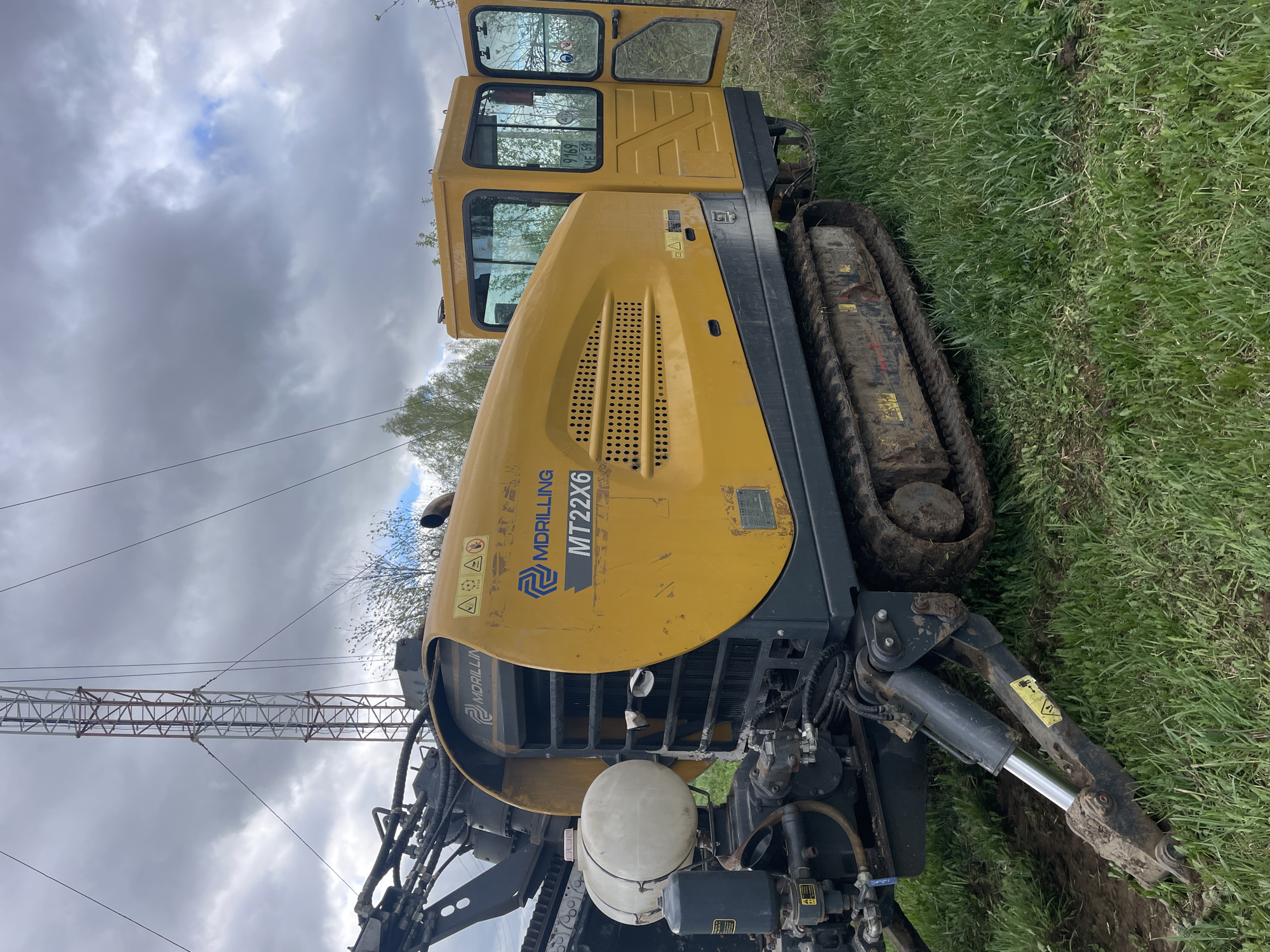 A yellow horizontal directional drilling rig, labeled "MDRILING MT22X6," sits in a grassy field. The rig has a tracked chassis and an open cab. A crane structure is visible in the background under a cloudy sky. The ground is a mix of grass and dirt, showing signs of recent activity.