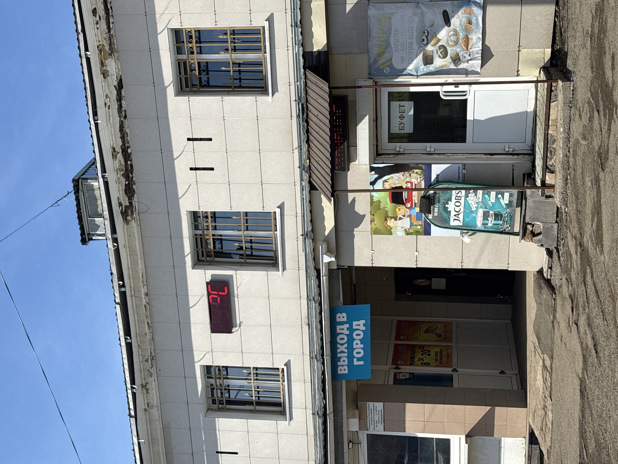 The image depicts the exterior of a building, likely a commercial or retail space, in Izhevsk, Russia. The building's façade is light-colored and tiled. It has several windows with ornate metal grilles. There is a digital display showing the temperature as 26 degrees Celsius. Various signs and advertisements are affixed to the building, including one for "JACOBS" coffee, a bakery, and a "BUFFET". Another sign indicates "EXIT TO CITY". There's an entrance below the "EXIT TO CITY" sign. The building appears to be adjacent to a paved street or parking area. The sky is blue.