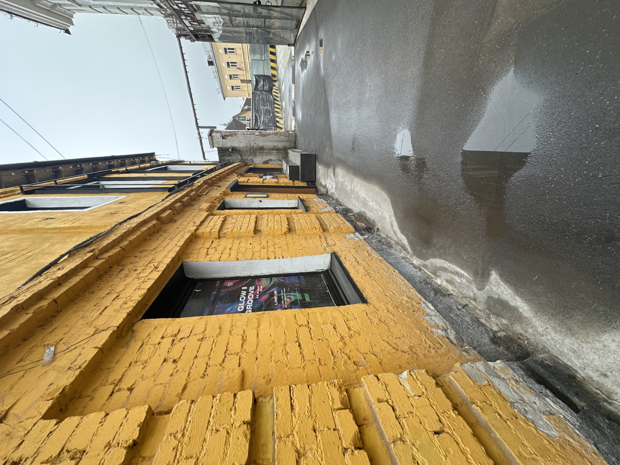 This image shows a view of an urban alleyway in Moscow, Russia. The camera is positioned at a low angle, looking up at a yellow brick building on the left. The building has several windows, one of which displays a poster advertising an event called "GLOW GROOVE" on "04 August." The alleyway is narrow, with a wet asphalt surface reflecting the sky. Other buildings are visible in the distance, appearing upside down due to the reflection in the puddles. The sky is overcast and gray. There are no people visible in the image.