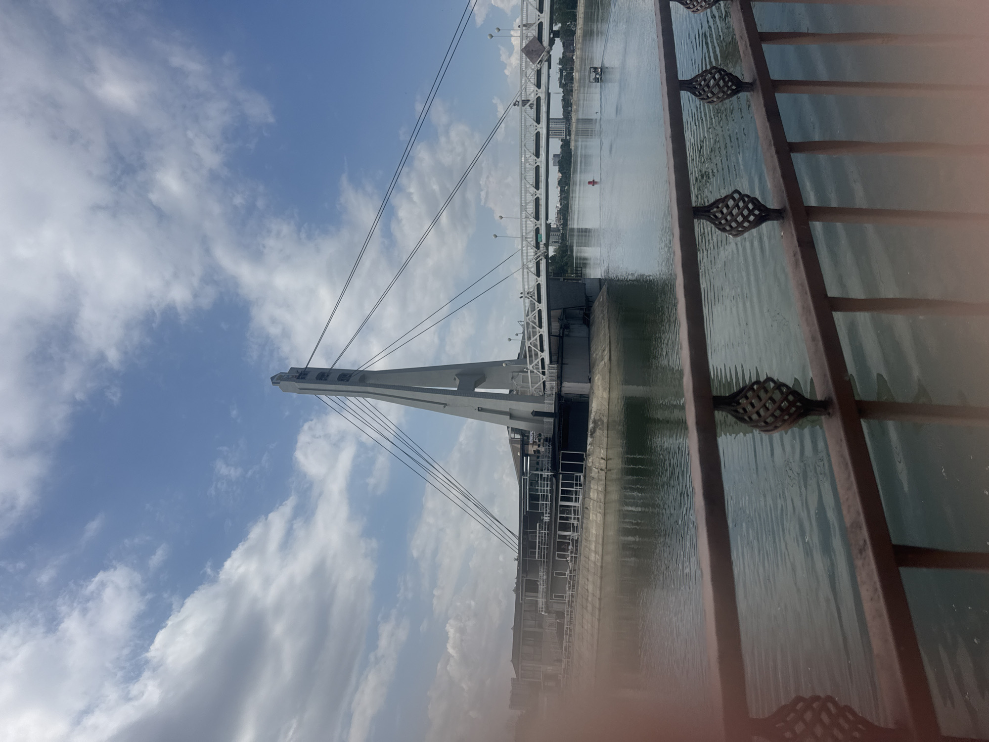 A wide shot captures a cable-stayed bridge spanning a body of water, likely a river or canal, in Krasnodar, Russia. The bridge is characterized by a tall, white, triangular pylon from which multiple cables extend to support the white pedestrian walkway. The walkway, illuminated by overhead lamps, is dotted with a few figures. On the left bank, a long, multi-story building with numerous windows is situated at the water's edge, adjacent to the bridge's foundation. The water reflects the sky and the structures. A decorative metal fence with intricate scrollwork runs across the foreground, partially obscuring the lower portion of the scene. The sky is a mix of blue with scattered white clouds. A red buoy and a small boat are visible in the water further down. The overall impression is one of a modern urban landscape integrated with a waterway.