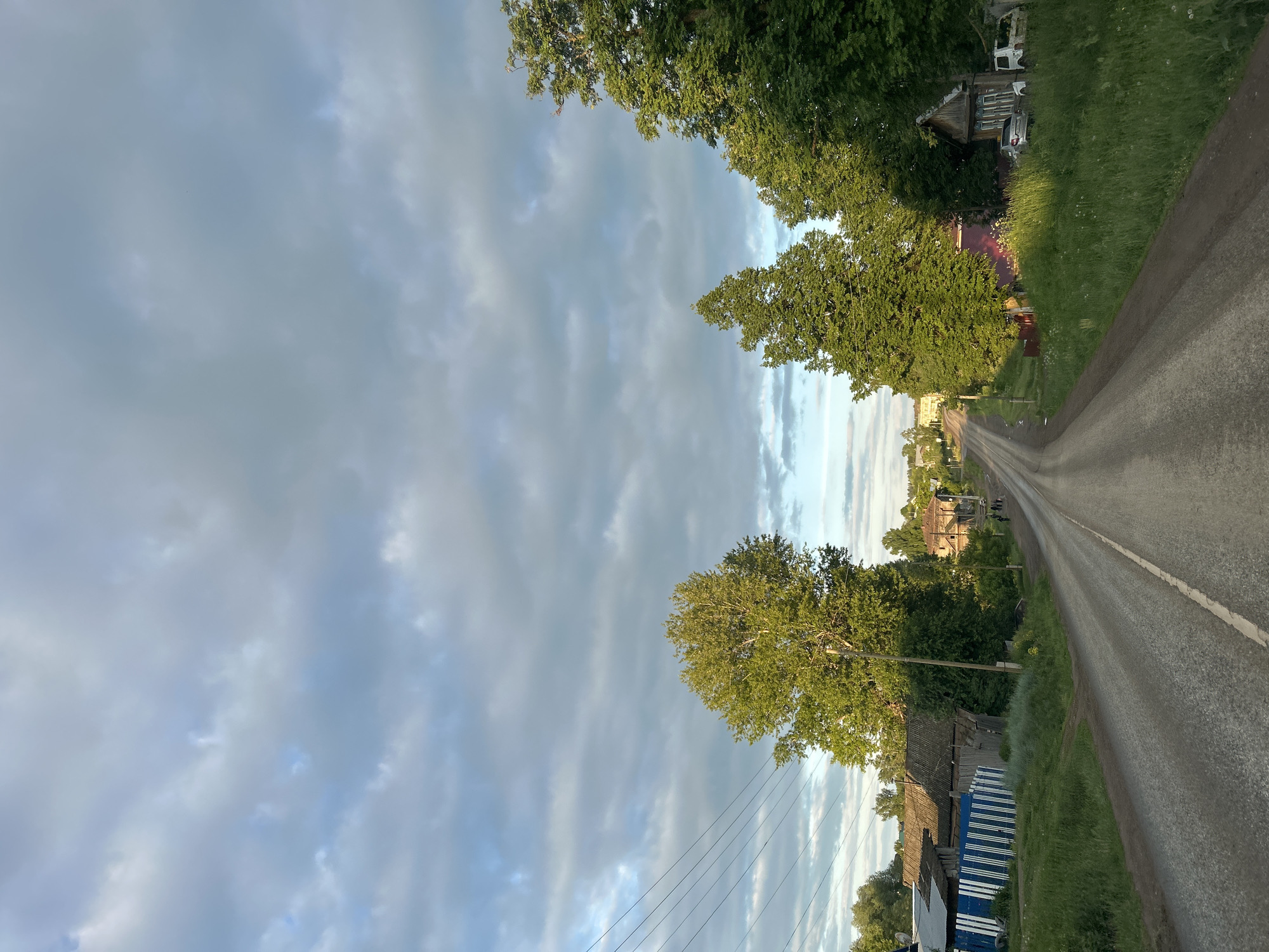 This image captures a long, straight rural road in Фоки, Russia, viewed from a slightly elevated perspective. The asphalt road, with a faint white line marking its right edge, extends into the distance, flanked by lush green trees and grassy verges. The sky above is partly cloudy, with a mix of light blue patches and soft grey clouds, suggesting late afternoon or early evening light, which casts a golden glow on some of the foliage.
On both sides of the road, traditional wooden houses and buildings are nestled among the trees. On the right, a white vehicle, possibly a van or truck, is partially visible behind trees and an old wooden structure. Further down, a silver car is parked near another house. On the left, a house with a dark roof is prominent, featuring a distinctive blue and white striped wooden fence in front of it. Utility poles with power lines run parallel to the road on this side. In the far distance, a small group of two to three indistinct figures can be seen on the road, appearing to be walking or standing. The scene conveys a tranquil, everyday atmosphere characteristic of a Russian village. No visible text is present.