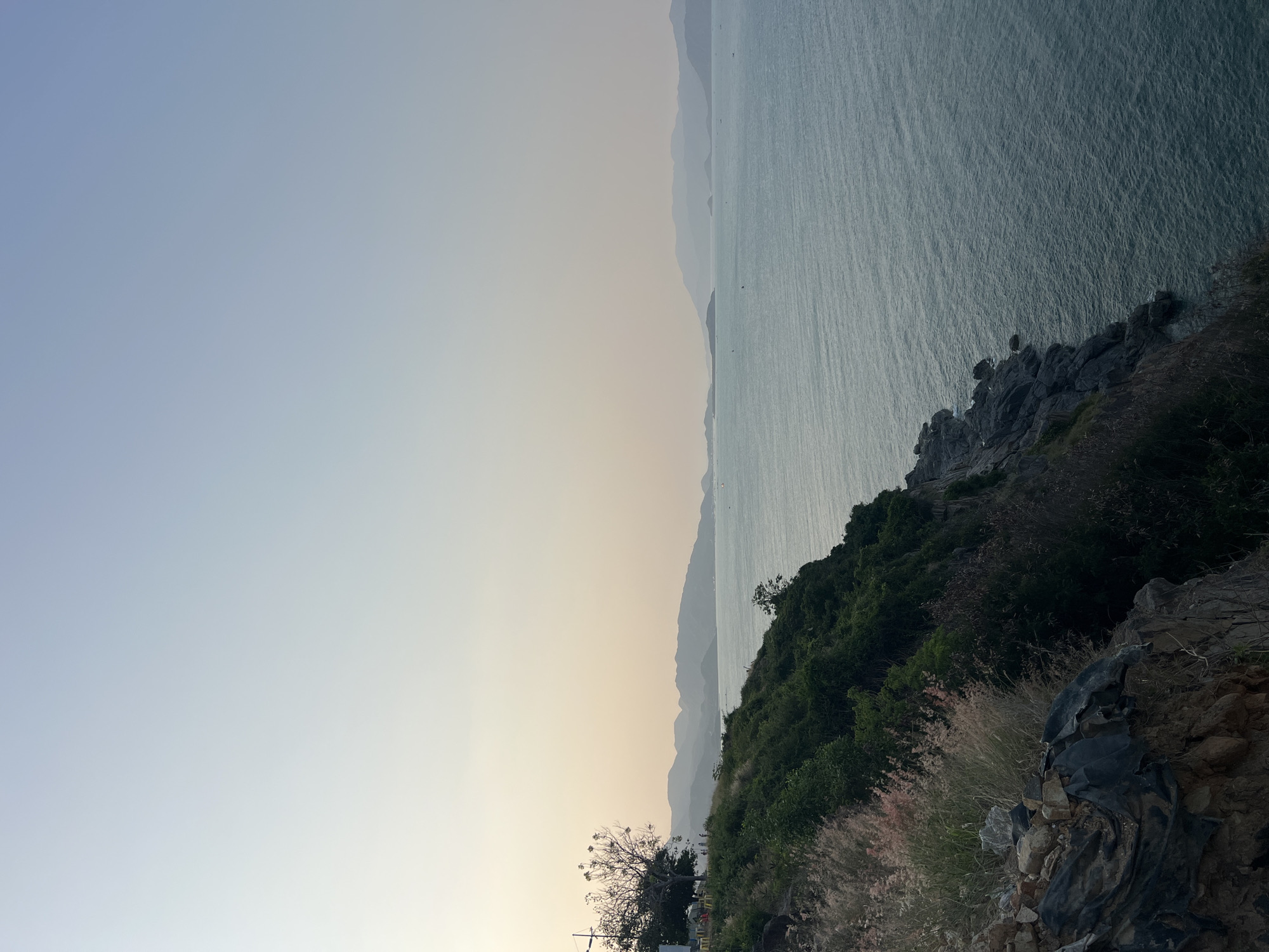 The image depicts a landscape in the Bac Natural Heritage Area Trang, Vietnam. The scene showcases a coastline with water extending into the horizon, distant mountains visible in the background. The foreground features vegetation, rocky terrain, and grassy areas. The sky shows a gradient from light blue to a pale yellow hue. There are no people visible in the image. The coastline is partly rocky.