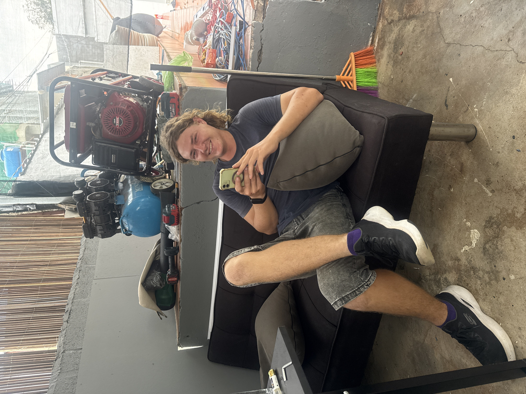A young man with curly brown hair and a beard is sitting on a black futon couch in an outdoor, urban setting. He is wearing a dark blue t-shirt, grey shorts, purple socks, and black and white athletic shoes. He is holding a light green smartphone in his hands and looking at it. The couch is positioned in front of a grey concrete wall, with a bamboo fence overhead. To the left of the couch, a small black table is visible with a glass of water and some tools. Behind the couch, there are two large pieces of machinery: a blue air compressor and a red generator. Further to the right, a broom with orange, green, and purple bristles leans against the wall. The ground is concrete and appears to be outdoors, possibly a workshop or storage area. The overall impression is one of a functional, perhaps slightly cluttered, outdoor workspace.