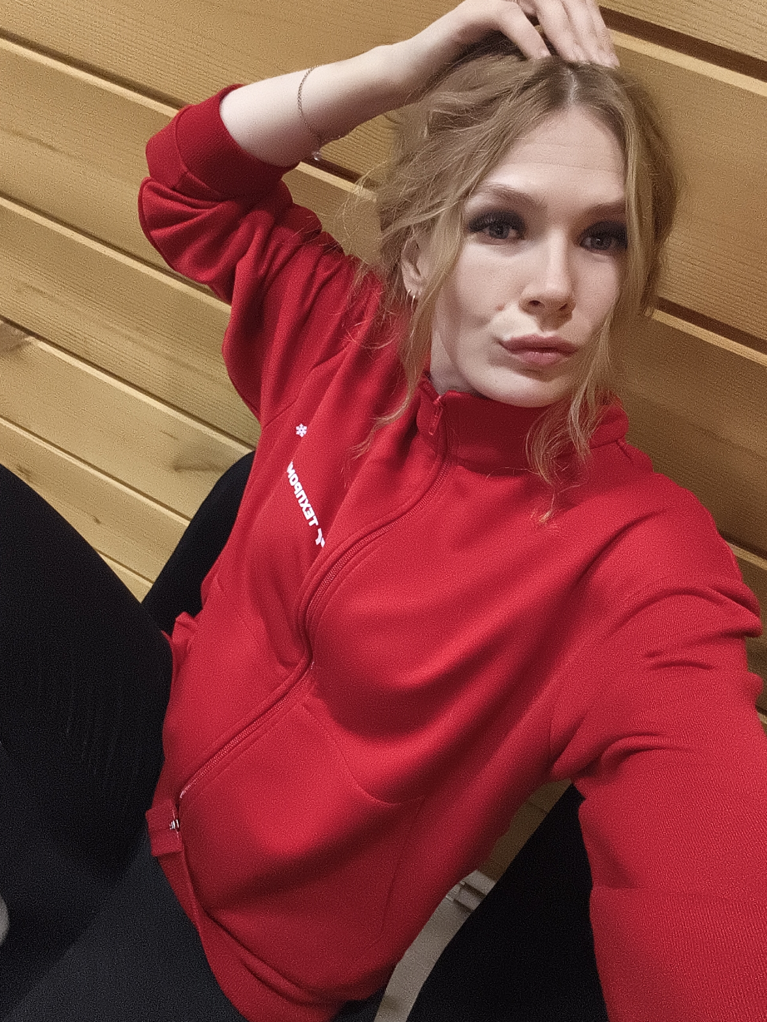 A young woman poses for a selfie indoors against a wood-paneled wall. She is seated, wearing a red zip-up jacket over black leggings. Her left hand is raised, touching her hair. The jacket has white lettering visible. The location is likely indoors, given the interior wood paneling. The city of Русский Вожой, Russia, is not discernible from the image's visual information.