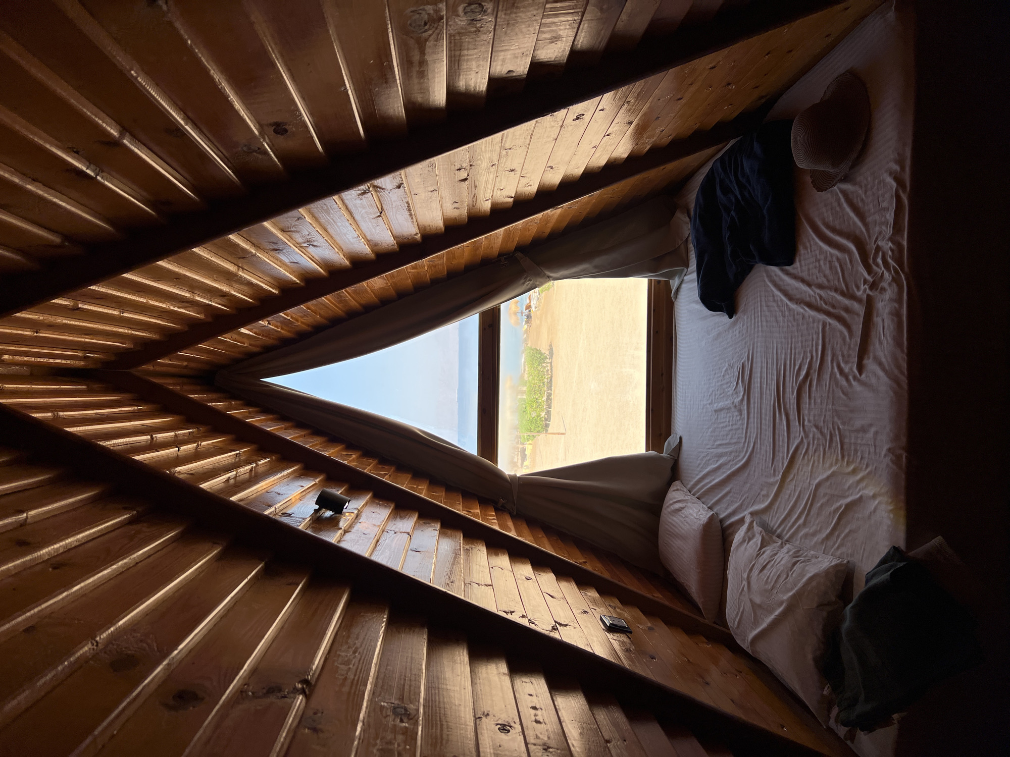 An interior shot of a triangular wooden room with a bed in the foreground, faces the window and the Red Sea. The wooden walls are made of horizontal planks, angled towards the apex of the triangle. The bed is covered in white sheets, with two pillows at the head. A dark blue blanket and a straw hat lie on the bed. Two small black wall lamps are visible on the wooden walls. Outside the window, a beach with some vegetation and umbrellas can be seen, with the sea beyond. The overall impression is of a cozy, rustic accommodation. The image is attributed to a location in Nuweiba, Egypt.