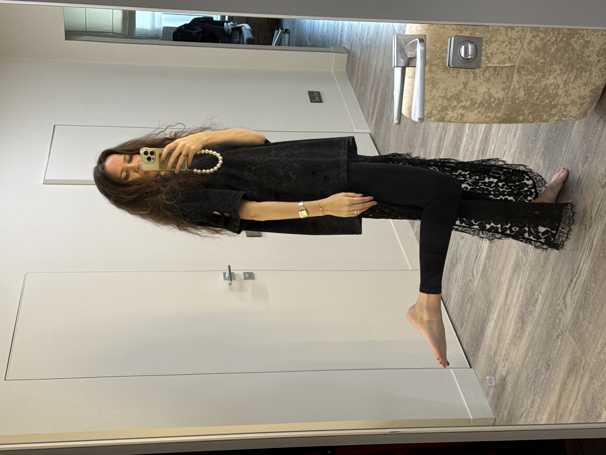 A woman is standing in a room, facing a mirror and holding up a smartphone to take a picture. The room has white walls, a light-colored wood floor, and a white door. A countertop with a faucet and sink is visible on the right side of the frame. The woman is wearing a black top, black leggings, and what appears to be a decorative black skirt overlay with a lace pattern. She has long, dark hair and is wearing a pearl necklace. A watch and bracelet are visible on her wrist. The setting is an interior space, possibly a dressing room or bathroom. There is no information available within the image to place the location within the city of Dolgoprudny, Russia.