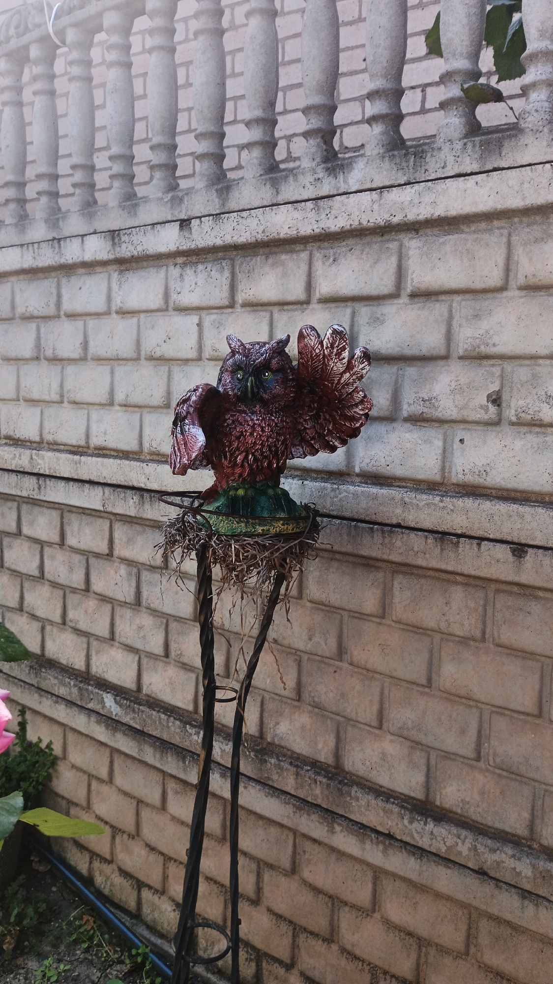 The image shows a decorative owl statue perched on a nest of twigs. The owl is made of a reddish-brown material with green eyes and is positioned with its wings spread as if in flight or defense. It stands on a decorative metal stand with twisted legs, adorned with a green base that supports the nest. The statue is set against a light-colored brick wall, topped by a concrete balustrade with decorative columns. The background suggests an outdoor setting, possibly a garden or courtyard in Kryvyi Rih, Ukraine, with a hint of greenery and a pink flower visible on the lower left. The overall scene is static, focusing on the statue as a decorative element.