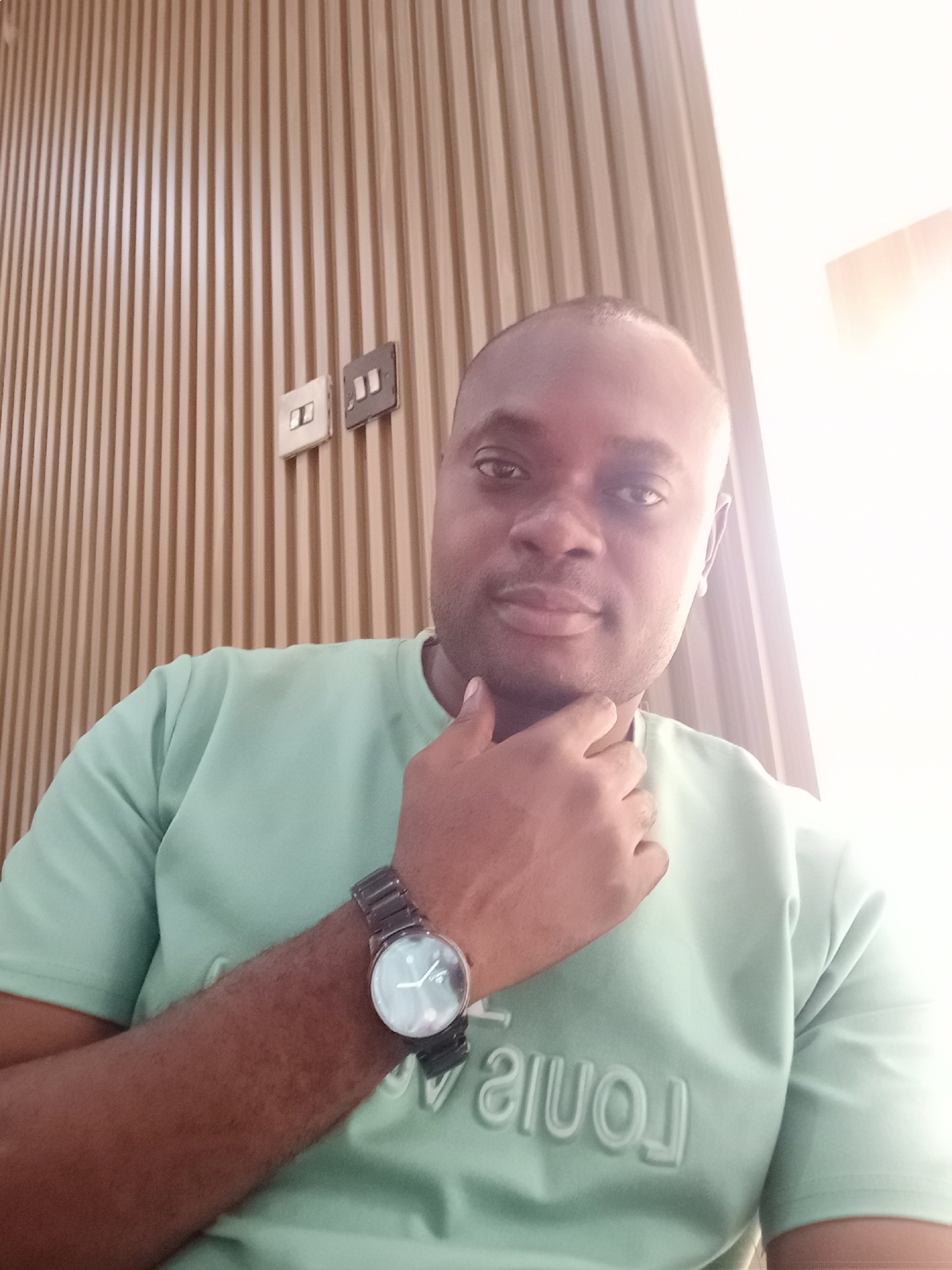 A medium, frontal shot of a dark-skinned man wearing a light green t-shirt and a dark-banded wristwatch. He is touching his chin with his right hand. The background consists of a light brown slatted wall with two electrical outlets. The location is likely indoors and in Gwarinpa, Nigeria.