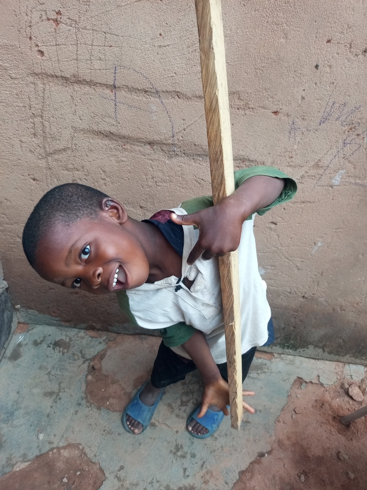 A young boy is shown outdoors, leaning against a rough, brown wall with some faint blue markings. He is wearing a white polo shirt with green sleeves and dark pants, along with blue sandals. He is holding a long, light-colored wooden plank vertically. The ground beneath him is a mix of cracked concrete and dirt. The image is shot from a high angle, looking down at the boy, who is smiling and looking directly at the camera. The setting appears to be in a residential or outdoor area in Tempure, Nigeria.