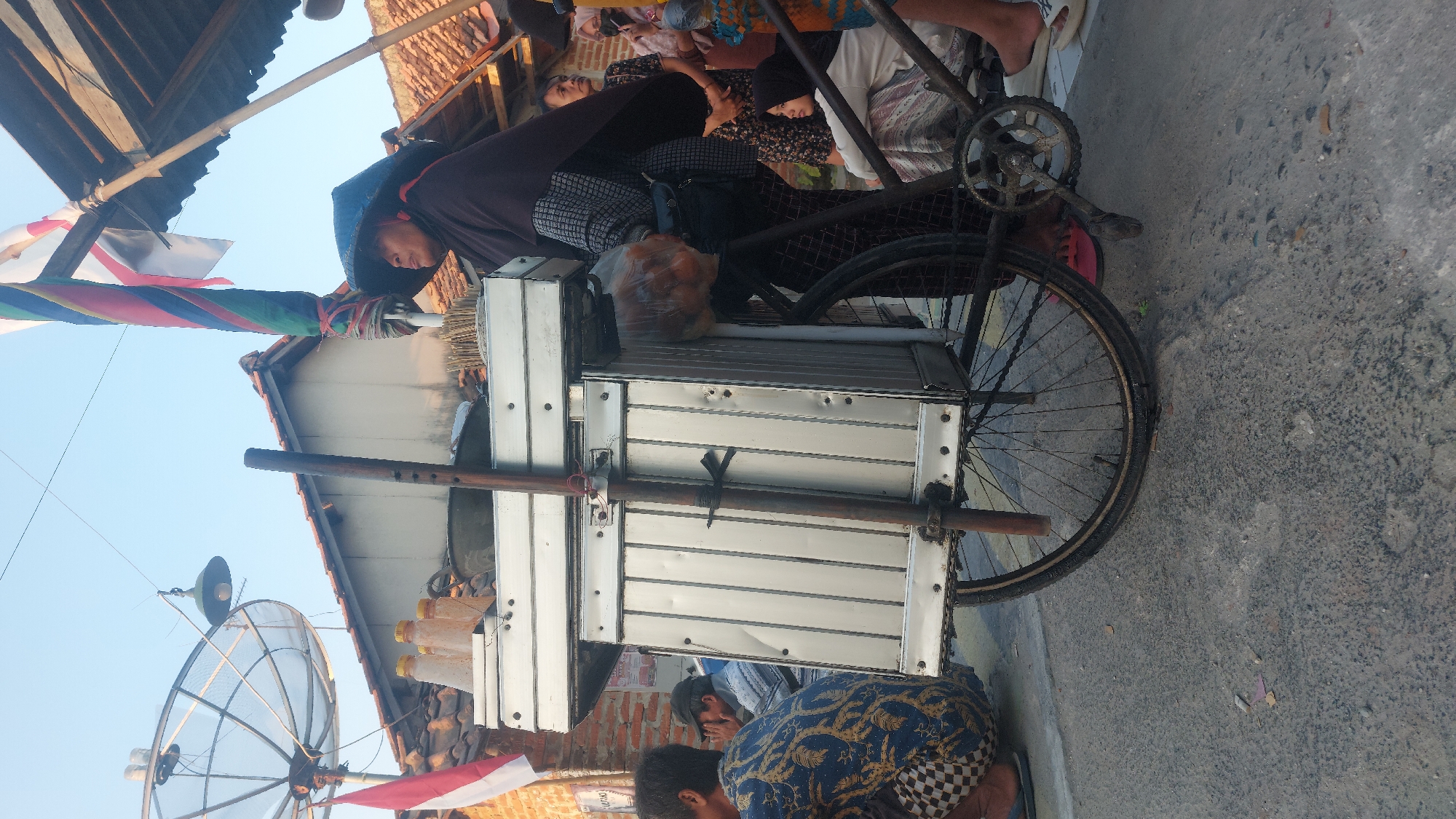 A street vendor, wearing a dark headscarf and a broad-brimmed hat, stands beside her mobile food cart in the city of Jatirogo, Indonesia. The cart, constructed of silver aluminum, is mounted on a bicycle frame. It is laden with several bottles of yellow liquid, presumably condiments or beverages, and a large plastic bag filled with spherical food items. A wooden pole, attached vertically to the cart, serves as a support.
In the foreground, a person in a patterned blue shirt and checkered pants is crouched on the ground. Behind the vendor, several other individuals are gathered, including a woman in a patterned dress and another wearing a dark hijab. A satellite dish and an Indonesian flag are visible in the background, atop the rooftops of several houses. The scene appears to be set on a paved street under a clear blue sky.